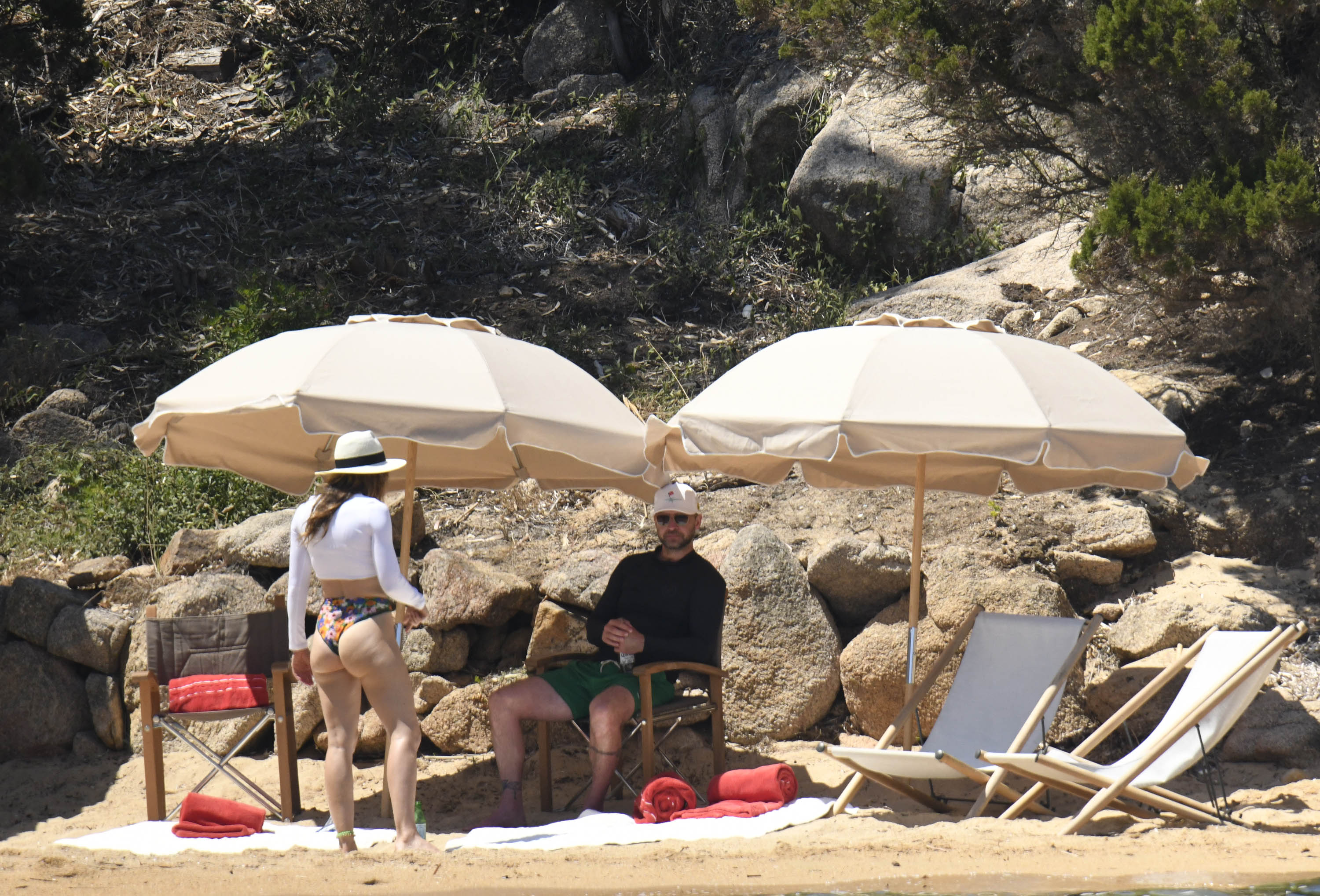 Jessica Biel stands beside Justin Timberlake during a beach outing. She wears a white long-sleeved crop top, colorful bikini bottoms, and a wide-brimmed hat, while Timberlake sits beneath a beach umbrella in a black long-sleeved top, green shorts, and a cap.