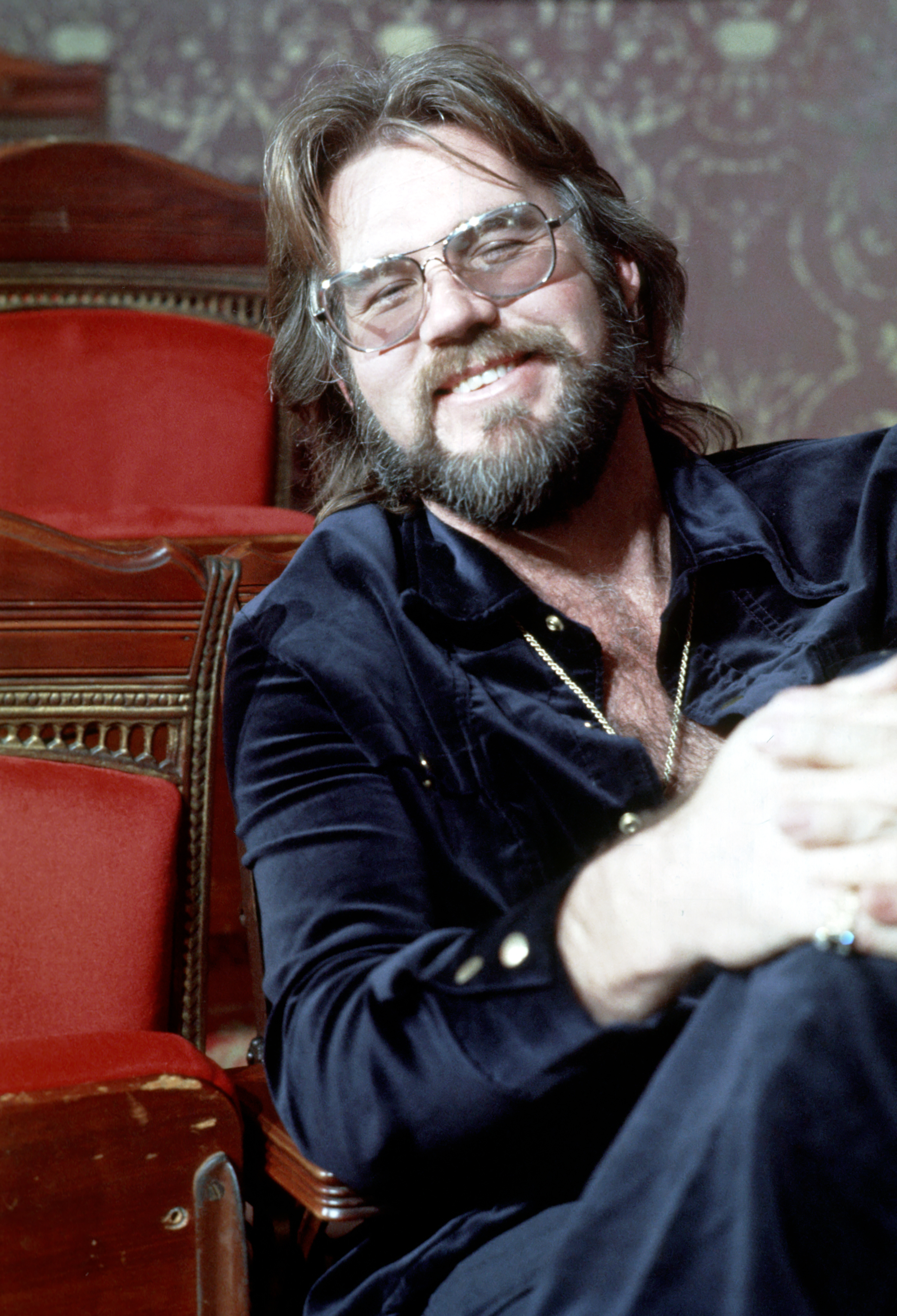 Kenny Rogers flashes an easy smile while seated in an ornate theater surrounded by red velvet seats in an archival portrait. Sporting his characteristic full beard, flowing dark hair, and wire-rimmed glasses, the musician relaxes in a navy velvet button-up shirt accented with gold buttons, paired with a gold chain necklace and rings. | Source: Getty Images