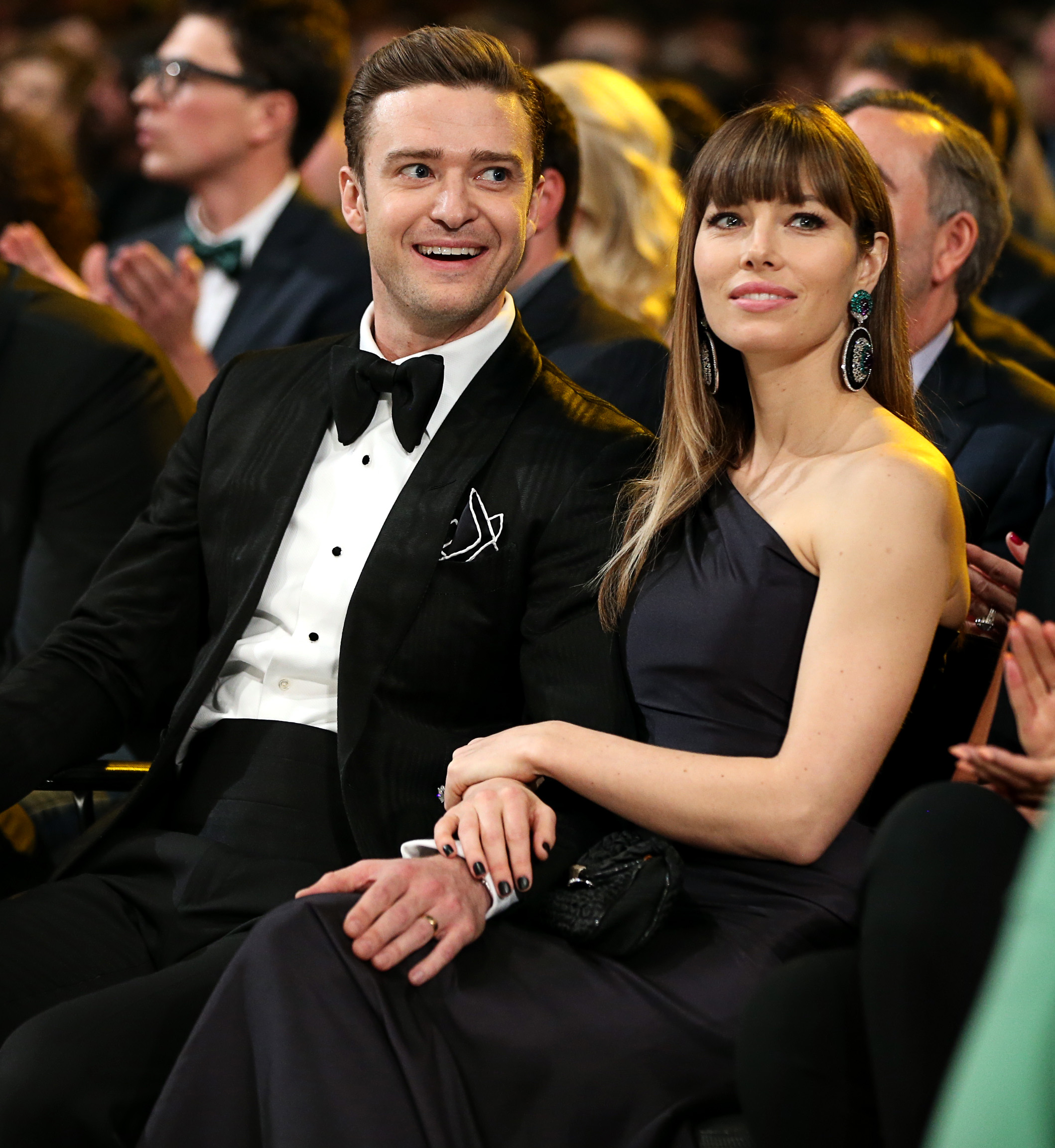 Justin Timberlake and Jessica Biel sit together at an awards ceremony, smiling as they watch the event. Timberlake wears a black tuxedo with a bow tie, while Biel stuns in a dark one-shoulder gown paired with statement earrings.