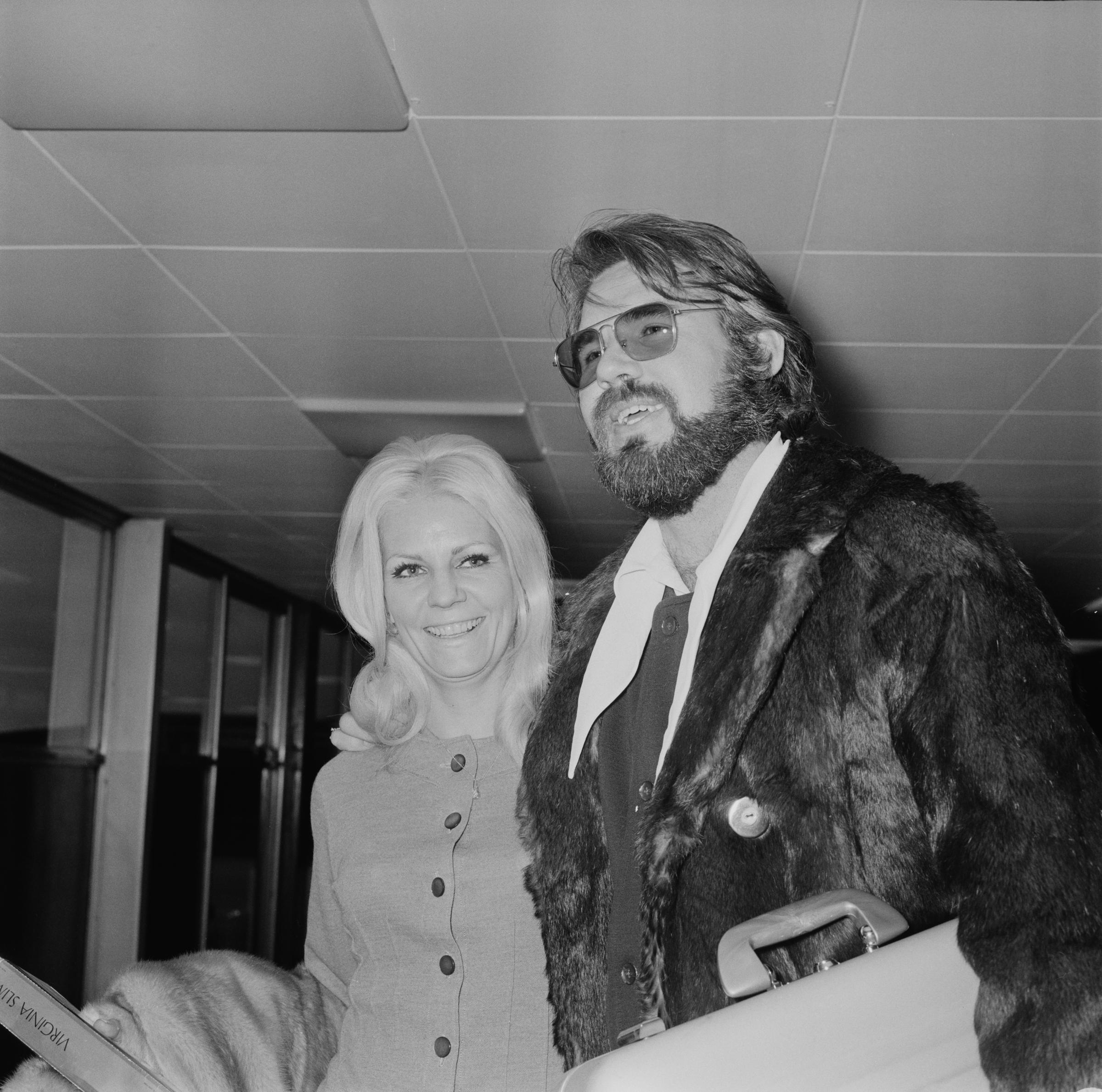 Kenny Rogers and his third wife, Margo Anderson, arrive at Heathrow Airport in London on March 24, 1970. The First Edition frontman sports tinted sunglasses, a full beard, a dark vest over a wide-collared white shirt, and a heavy fur coat while carrying luggage, as blonde Margo smiles alongside him in a tailored, buttoned jacket. | Source: Getty Images