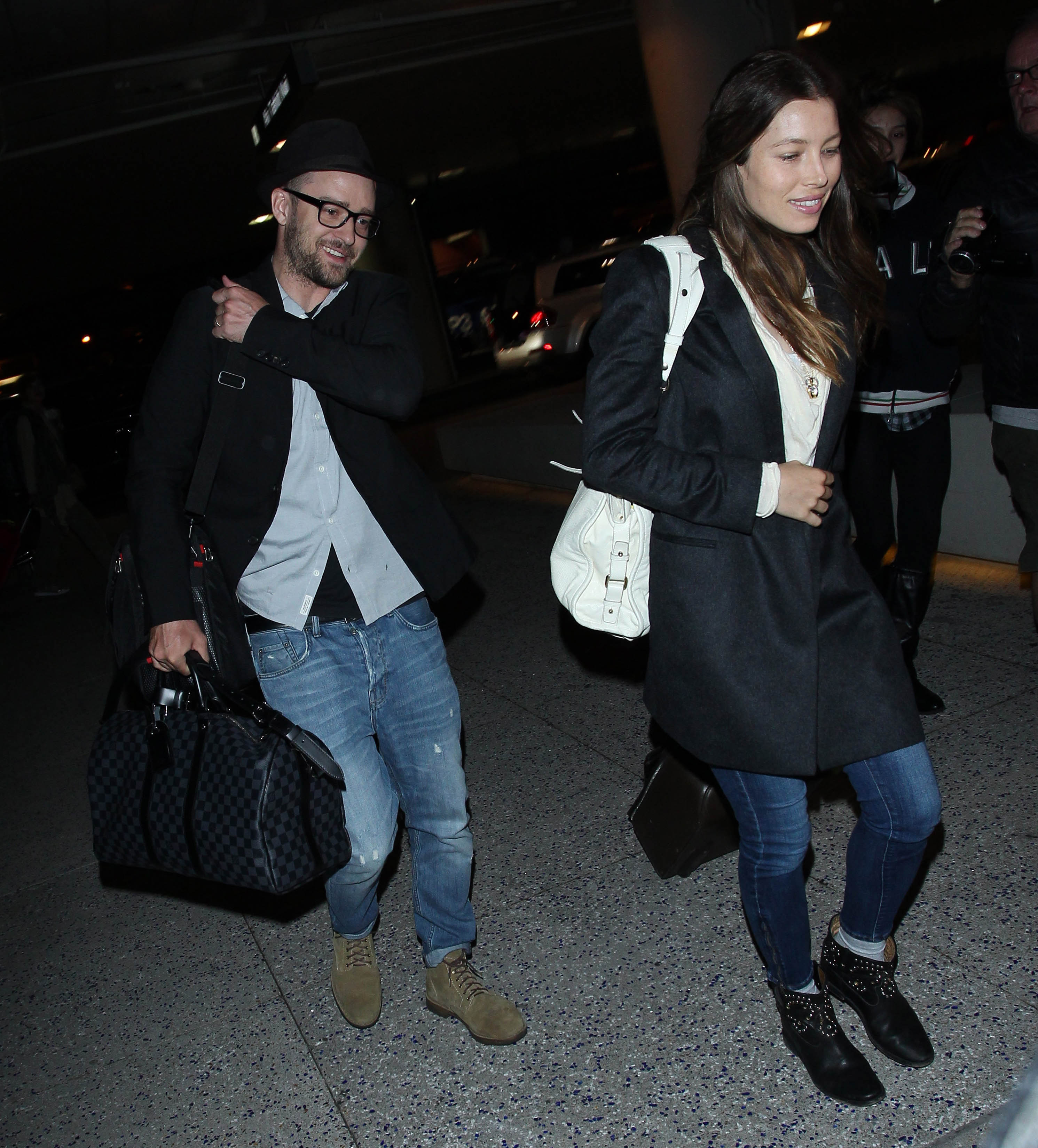 Justin Timberlake and Jessica Biel arrive at an event together, carrying bags as they walk through a parking area. Timberlake wears a dark blazer, glasses, and a black hat, while Biel pairs a dark coat with jeans, ankle boots, and a white backpack.