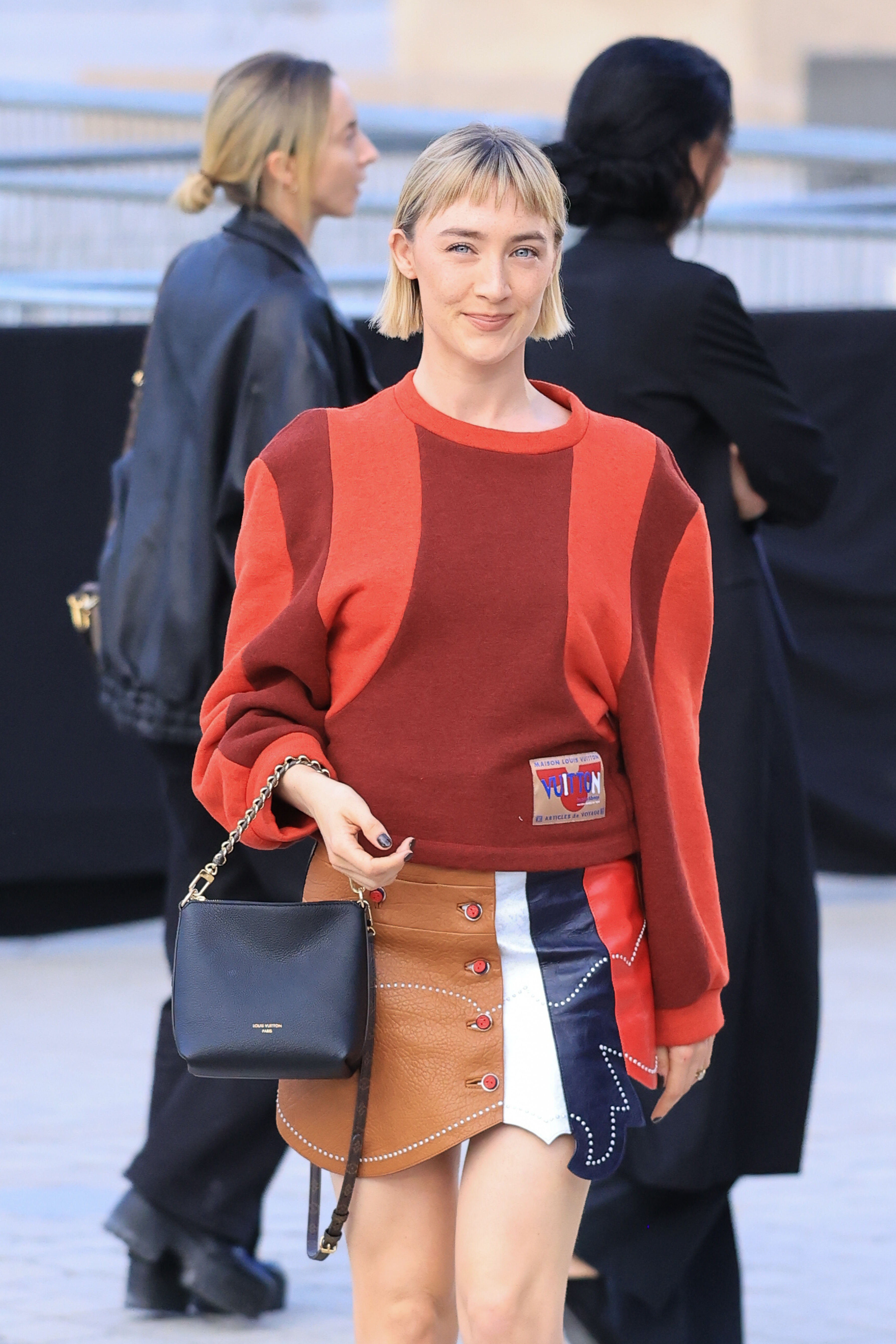 Saoirse Ronan attends the Louis Vuitton Womenswear Spring/Summer 2027 show as part of Paris Fashion Week on October 06, 2026, in Paris, France. She wore a red-and-burgundy color-blocked top paired with an asymmetrical patchwork mini skirt in tan, white, navy, and red. A structured black Louis Vuitton handbag and her short blonde bob gave the playful look a polished finish. | Source: Getty Images