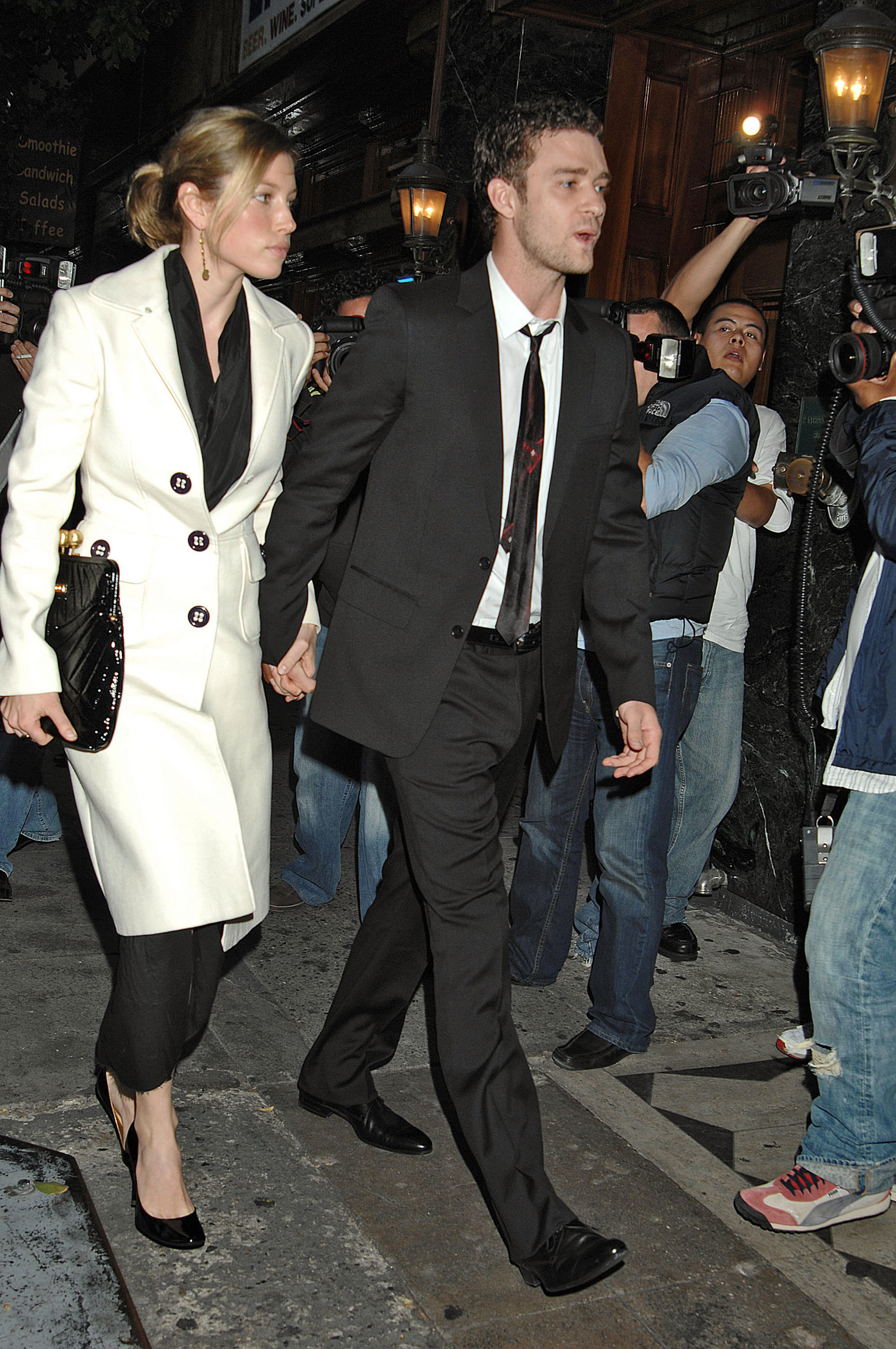 essica Biel and Justin Timberlake walk hand in hand through Los Angeles, with Biel wearing a white coat over a black outfit and Timberlake sporting a dark suit and tie. | Source: Getty Images