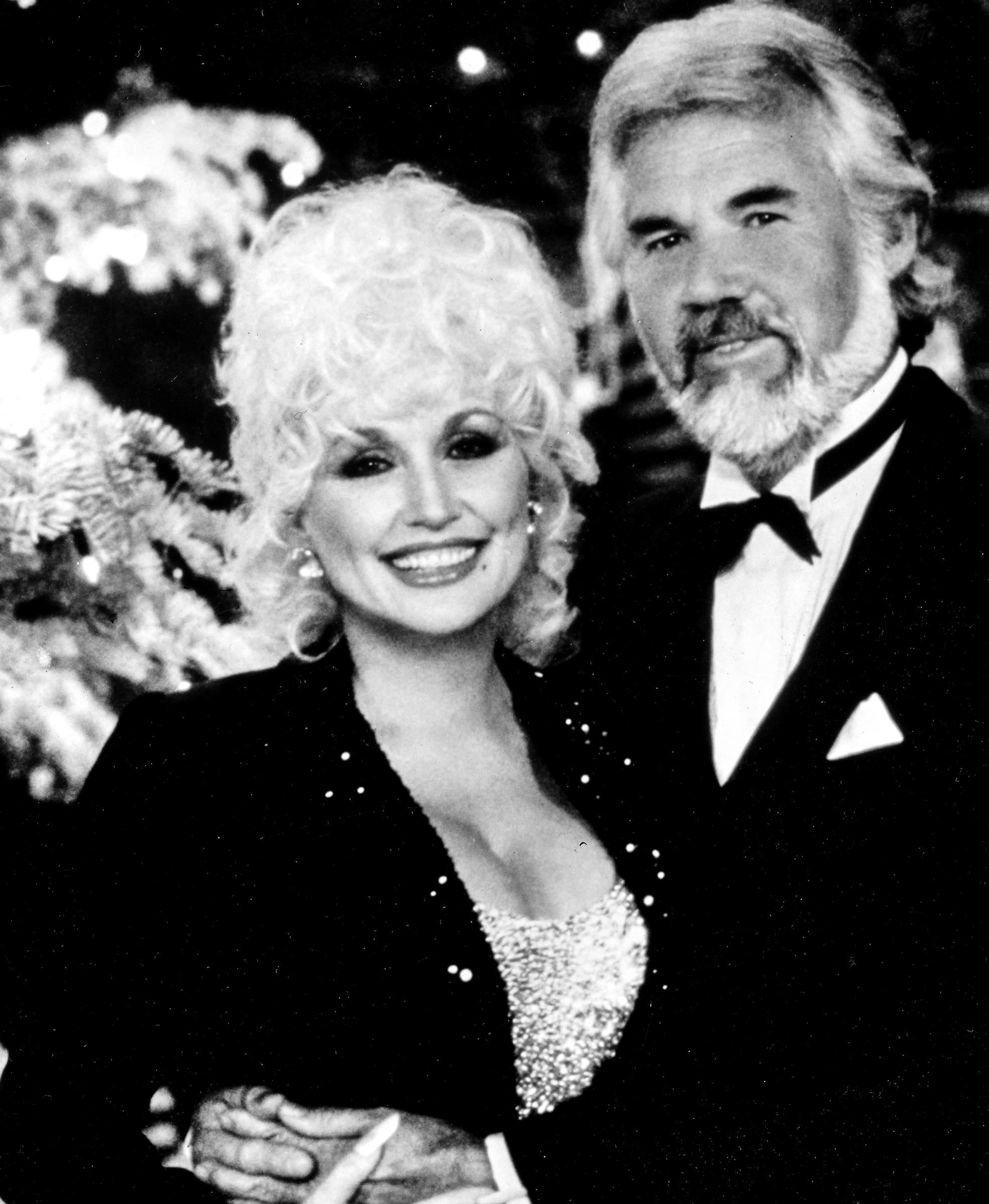 Dolly Parton and Kenny Rogers pose together in formal holiday attire in February 6, 1980. Dolly smiles warmly with her signature curls, wearing a dark jacket accented with subtle sparkle over a shimmering sequined top, while Kenny embraces her from behind in a classic black tuxedo, bow tie, and pocket square in front of a decorated Christmas tree. | Source: Getty Images