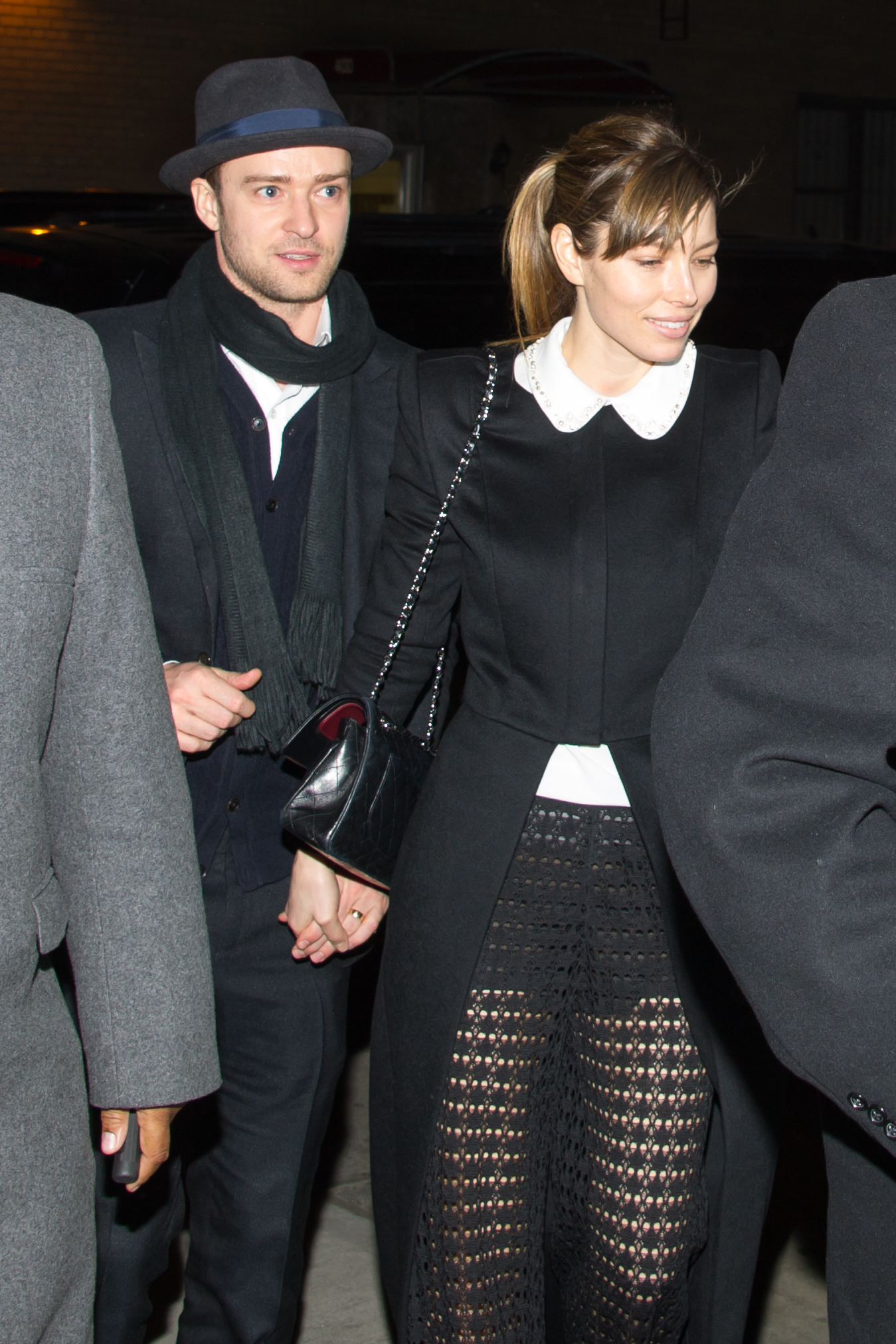 Justin Timberlake and Jessica Biel hold hands while walking outdoors at night. Timberlake wears a dark coat, scarf, and fedora, while Biel sports a black coat with a white Peter Pan collar and a patterned skirt.