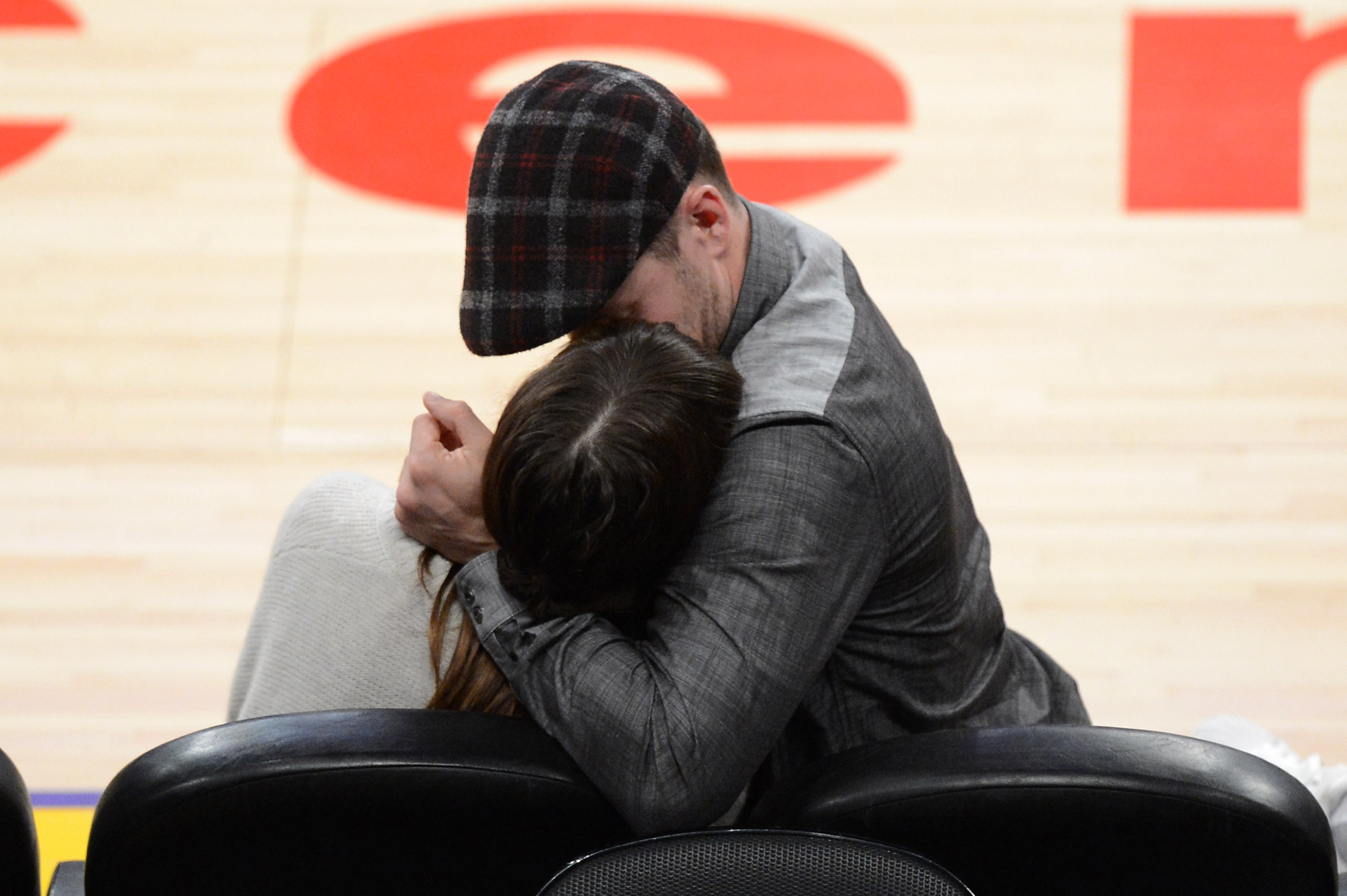 Justin Timberlake embraces Jessica Biel as they sit courtside at a basketball game, sharing a tender moment while surrounded by the arena's bright court lights. Timberlake wears a gray jacket and plaid flat cap. | Source: Getty Images