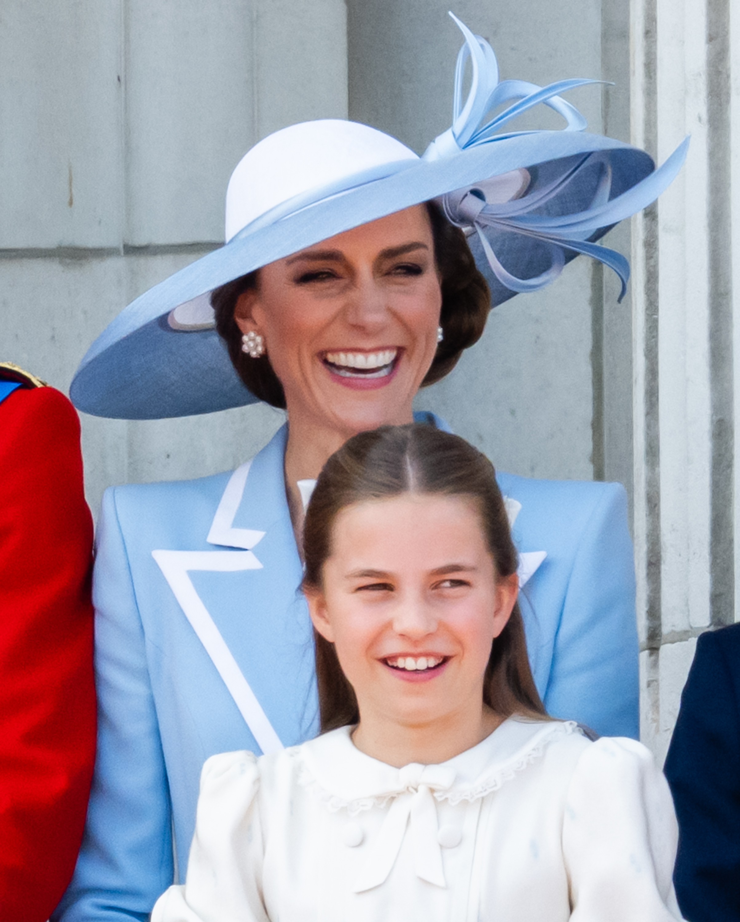 Princess Charlotte of Wales and Catherine, Princess of Wales share a joyful laugh on the balcony of Buckingham Palace while watching the RAF flypast during Trooping the Colour on June 13, 2026, in London, England. Charlotte smiles brightly in an ivory dress detailed with a ruffled lace collar and bow at the neckline, standing in front of Catherine, who wears a powder-blue tailored coat dress featuring crisp white lapel accents, pearl cluster earrings, and an architectural Philip Treacy hat trimmed with sculpted ribbon loops. | Source: Getty Images