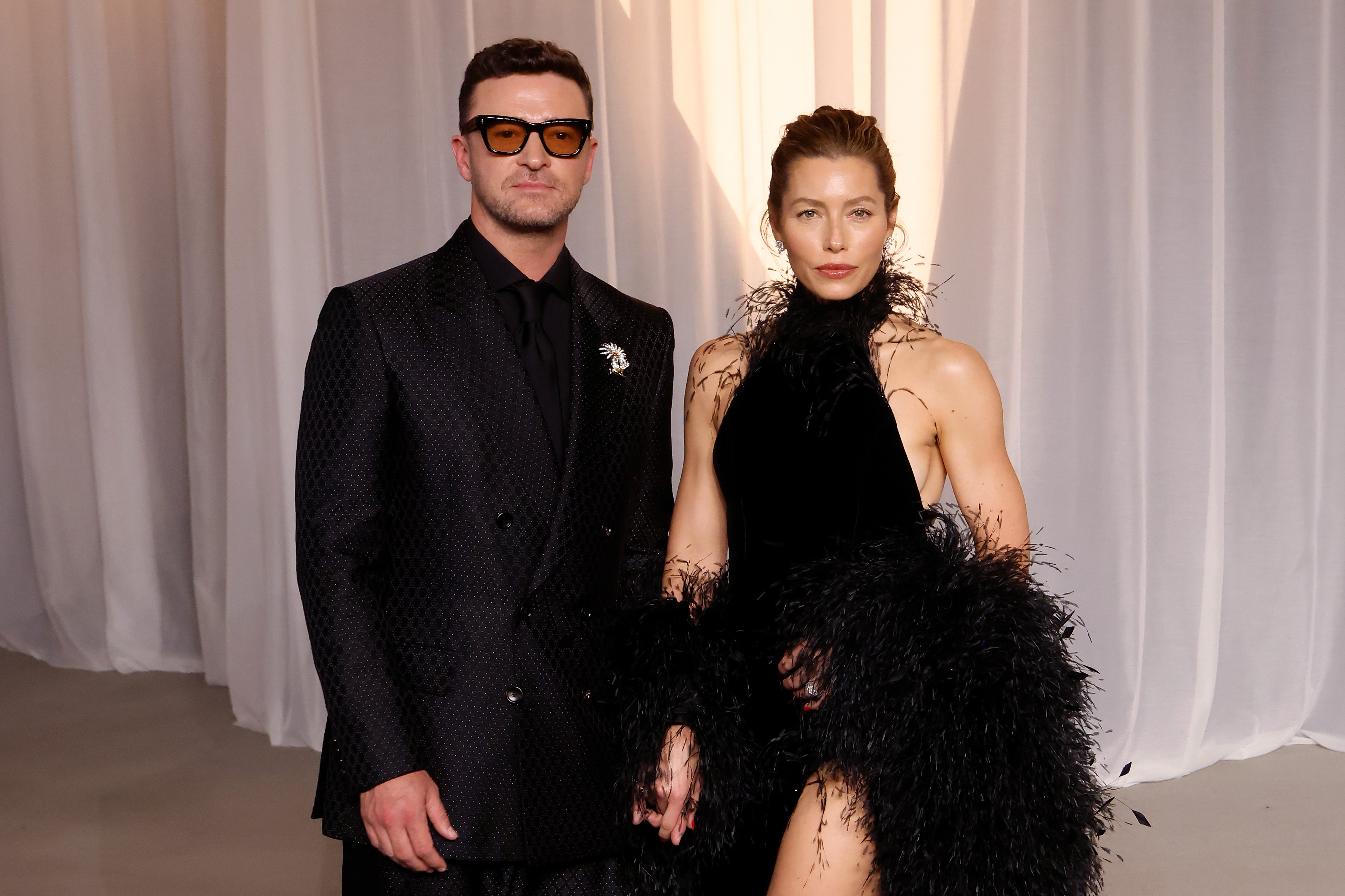 Jessica Biel and Justin Timberlake pose side by side in coordinated black ensembles. Biel's sleeveless gown features striking feathered details and a high slit, while Timberlake wears a black patterned suit, a black shirt and tie, and tinted glasses.