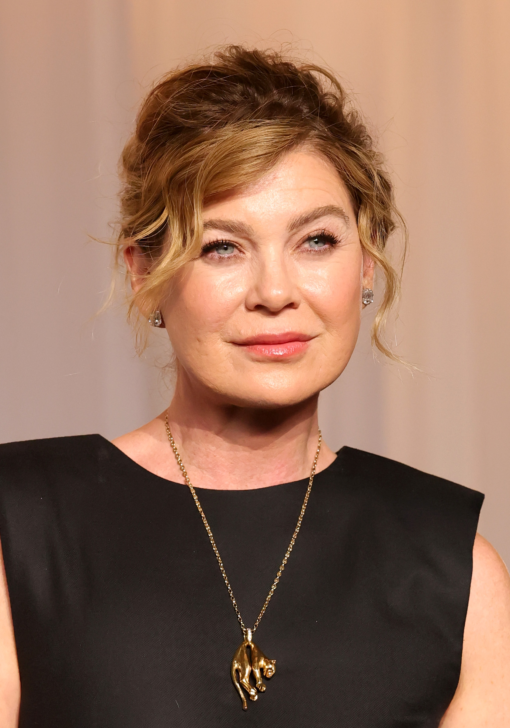 Ellen Pompeo poses for a close-up portrait with her hair swept into a loose updo, pairing soft pink makeup with stud earrings and a long gold pendant necklace.