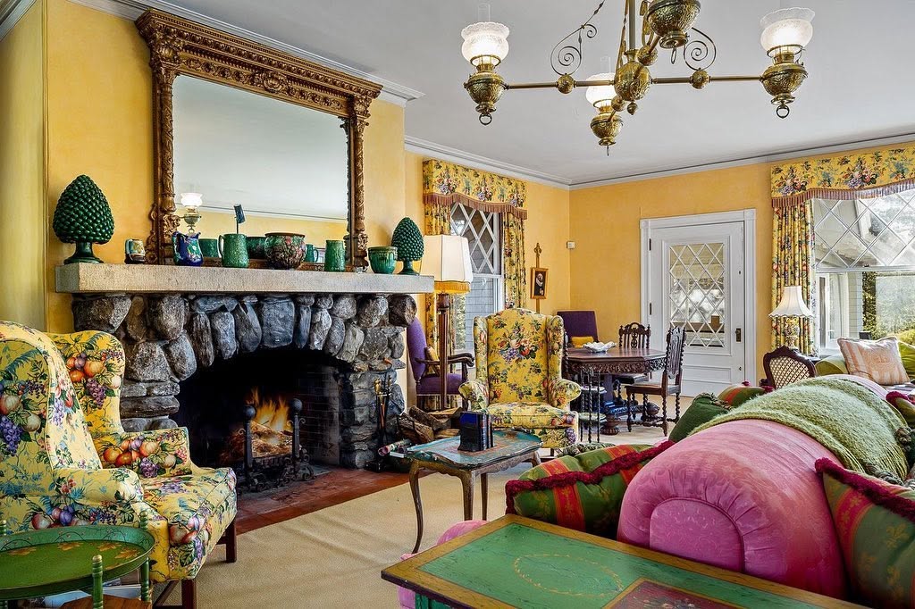 A colorful sitting room pairs a large stone fireplace with floral armchairs, bright furnishings, and an ornate mirror above the mantel. | Source: Facebook/LegacySIR