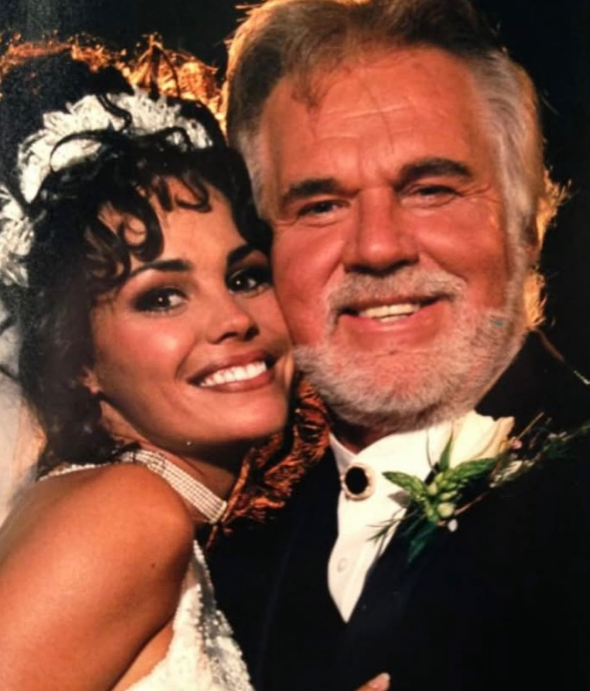 Kenny Rogers and his fifth wife, Wanda Miller, beam closely together in a romantic wedding portrait from their ceremony at Kenny’s ranch in Athens, Georgia, in 1997. A radiant Wanda wears an embellished bridal gown, headpiece, and pearl choker necklace as she leans against Kenny, who smiles in a black tuxedo accented with a bolo tie and floral boutonnière.