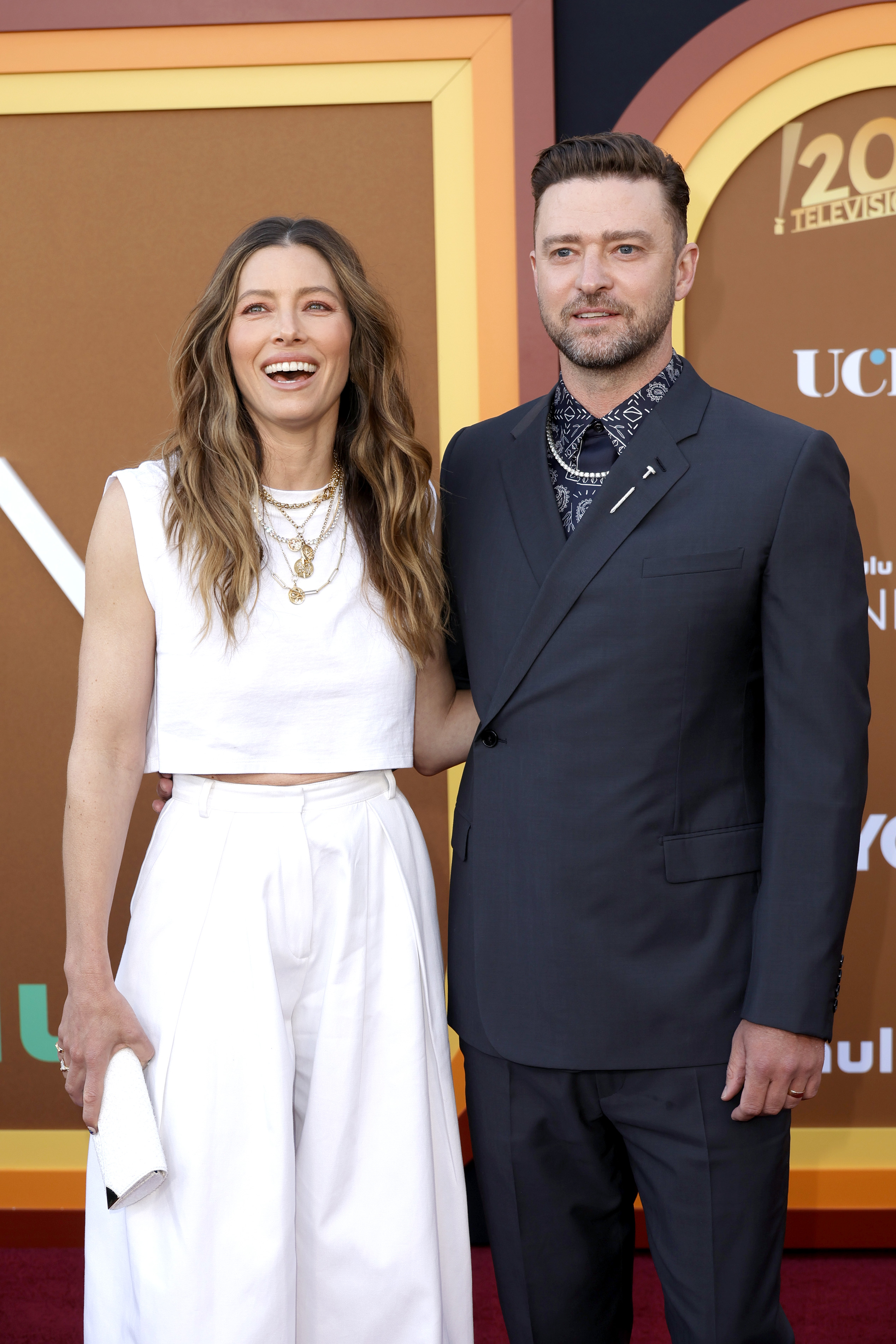 Justin Timberlake and Jessica Biel pose together on the red carpet at the 2024 Primetime Emmy Awards. Biel laughs in a white sleeveless crop top and matching wide-leg trousers, accessorized with layered gold necklaces, while Timberlake wears a navy suit with a patterned shirt and tie.