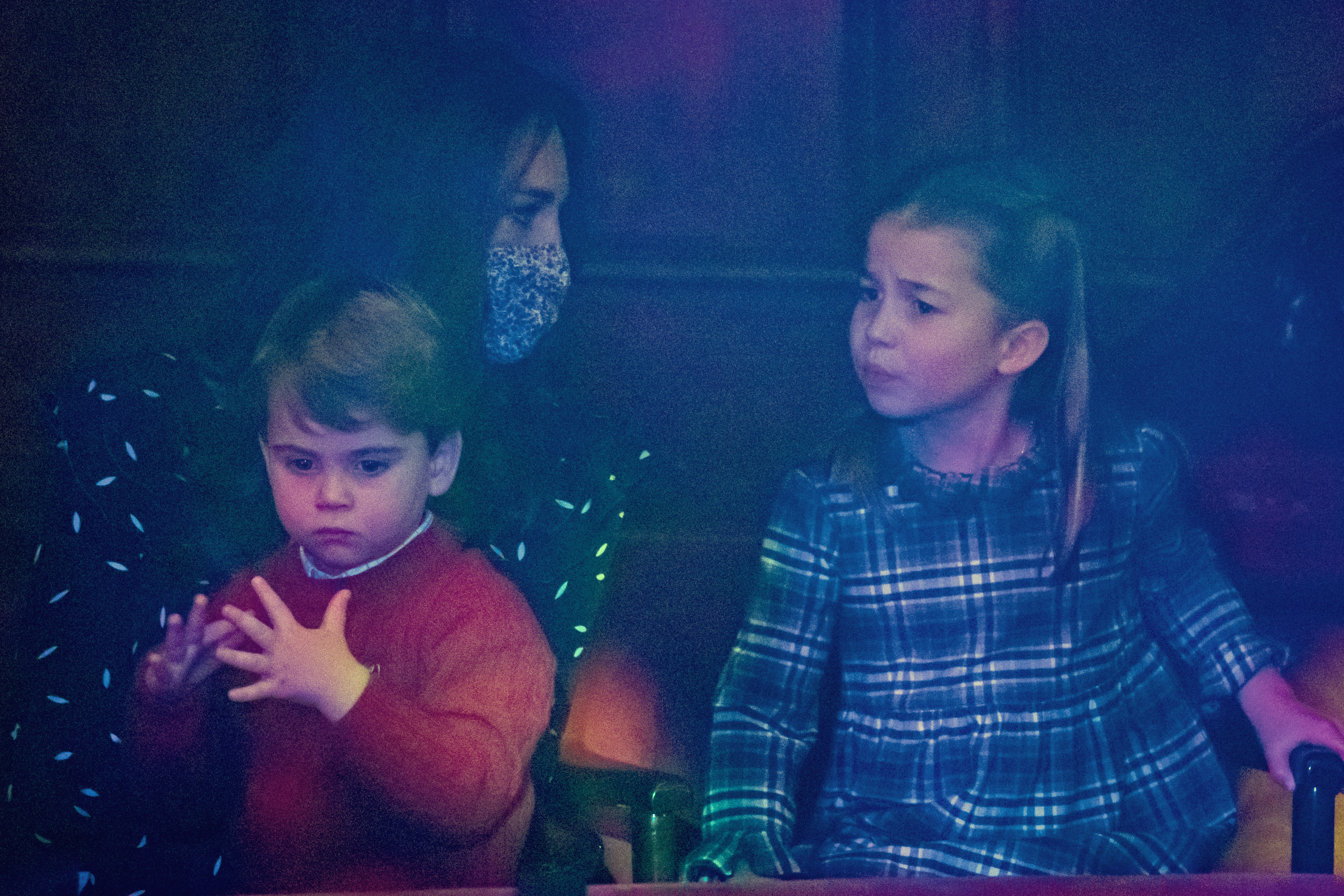 Princess Charlotte and Prince Louis watch from the audience during a special pantomime performance at the London Palladium Theatre on December 11, 2020, in London, England, held to thank key workers and their families for pandemic efforts. Charlotte sits attentively in a blue-and-green tartan dress, beside Louis in a cozy red crewneck sweater over a collared shirt, while their mother Catherine, Duchess of Cambridge looks on wearing a printed dress and a protective face mask. | Source: Getty Images