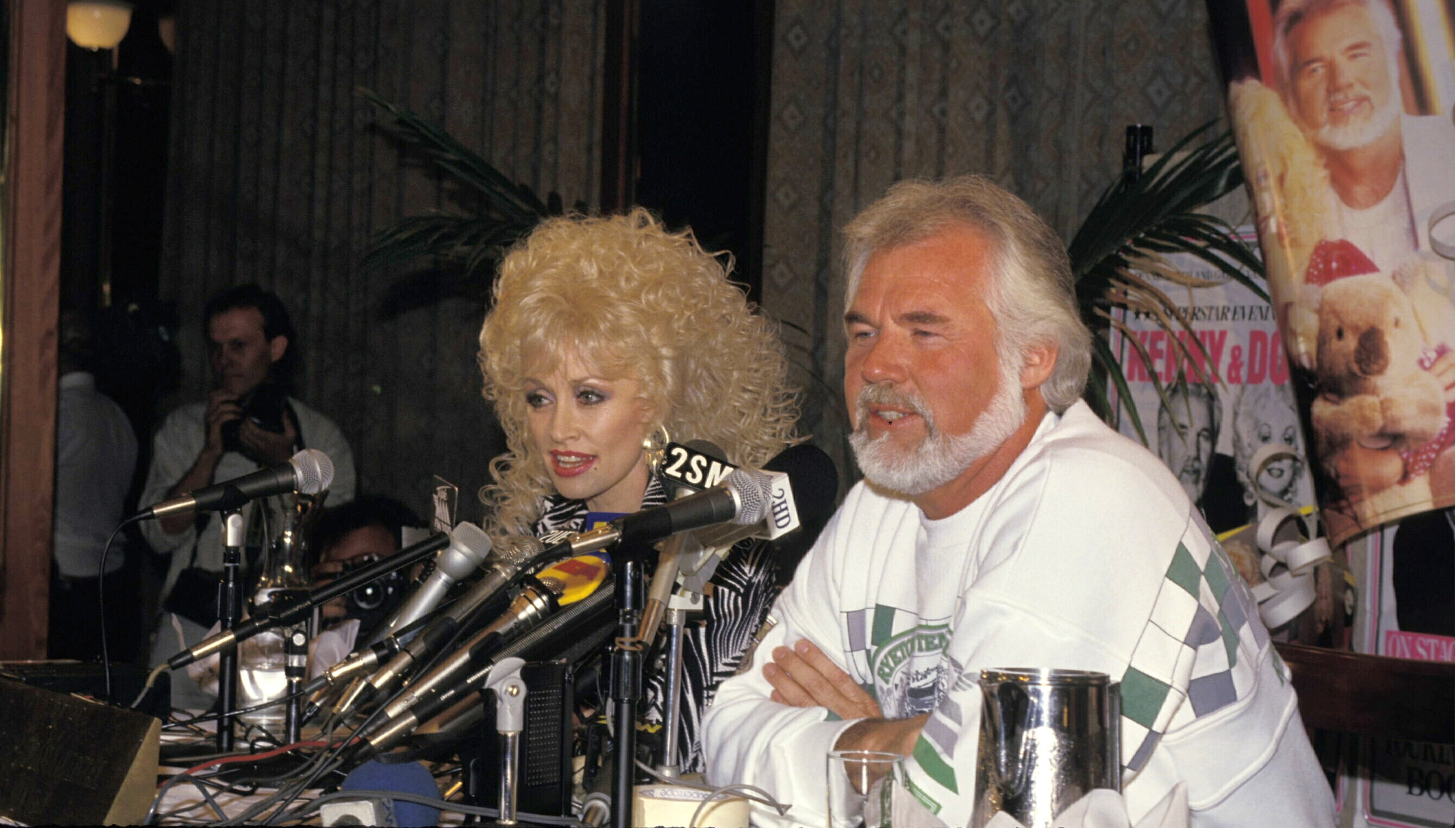 Country music icons Dolly Parton and Kenny Rogers sit side by side during a joint press conference in 1987. Facing a table filled with media microphones, Dolly speaks with her signature voluminous blonde curls in a graphic patterned top, while Kenny sits beside her in a casual white patterned sweatshirt, listening attentively. | Source: Getty Images