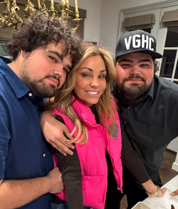 Wanda Miller smiles warmly as she is embraced by her adult twin sons, Justin and Jordan Rogers, in a relaxed family photo. Wanda wears a bright pink quilted vest over a dark long-sleeve top, flanked by her boys—one with curly hair in a blue collared shirt leaning in close, and the other smiling beside her in a black shirt and a baseball cap.