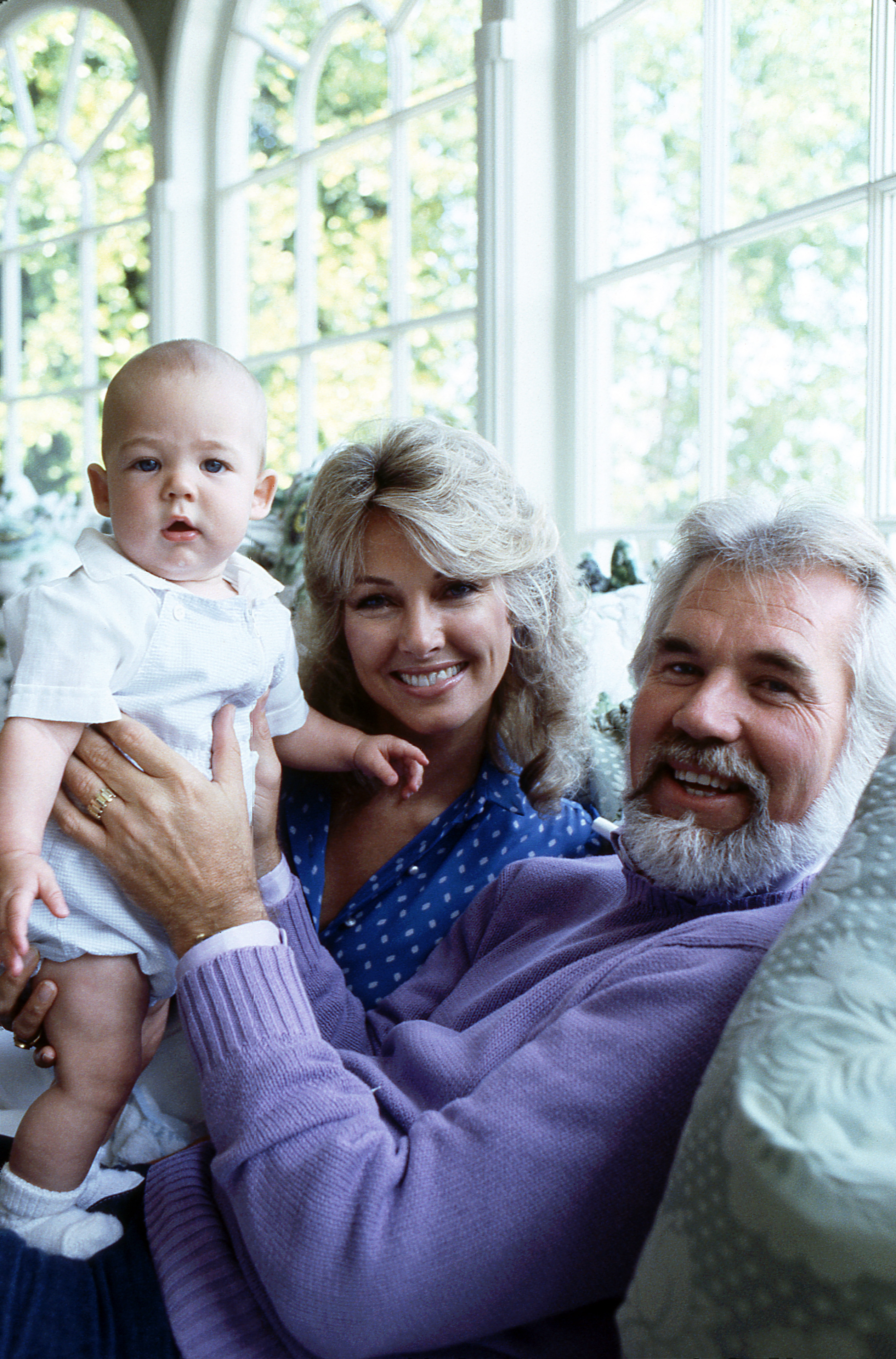 Kenny Rogers and his fourth wife, actress Marianne Gordon, pose with their infant son, Christopher Cody Rogers, during an appearance on "20/20" in 1982. Kenny smiles warmly in a lavender knit sweater as he cradles baby Christopher, dressed in a white romper, while Marianne beams beside them in a blue polka-dot blouse in front of large, sunlit windows.