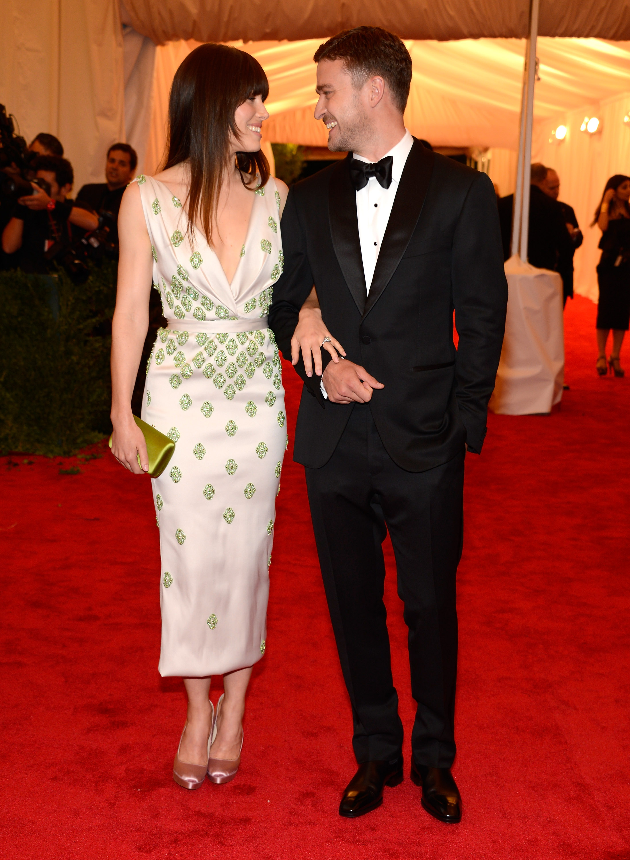 Jessica Biel and Justin Timberlake share a smile while walking arm in arm at the Met Gala. Biel wears a white gown adorned with green embellishments and carries a matching clutch, while Timberlake looks sharp in a classic black tuxedo. | Source: Getty Images