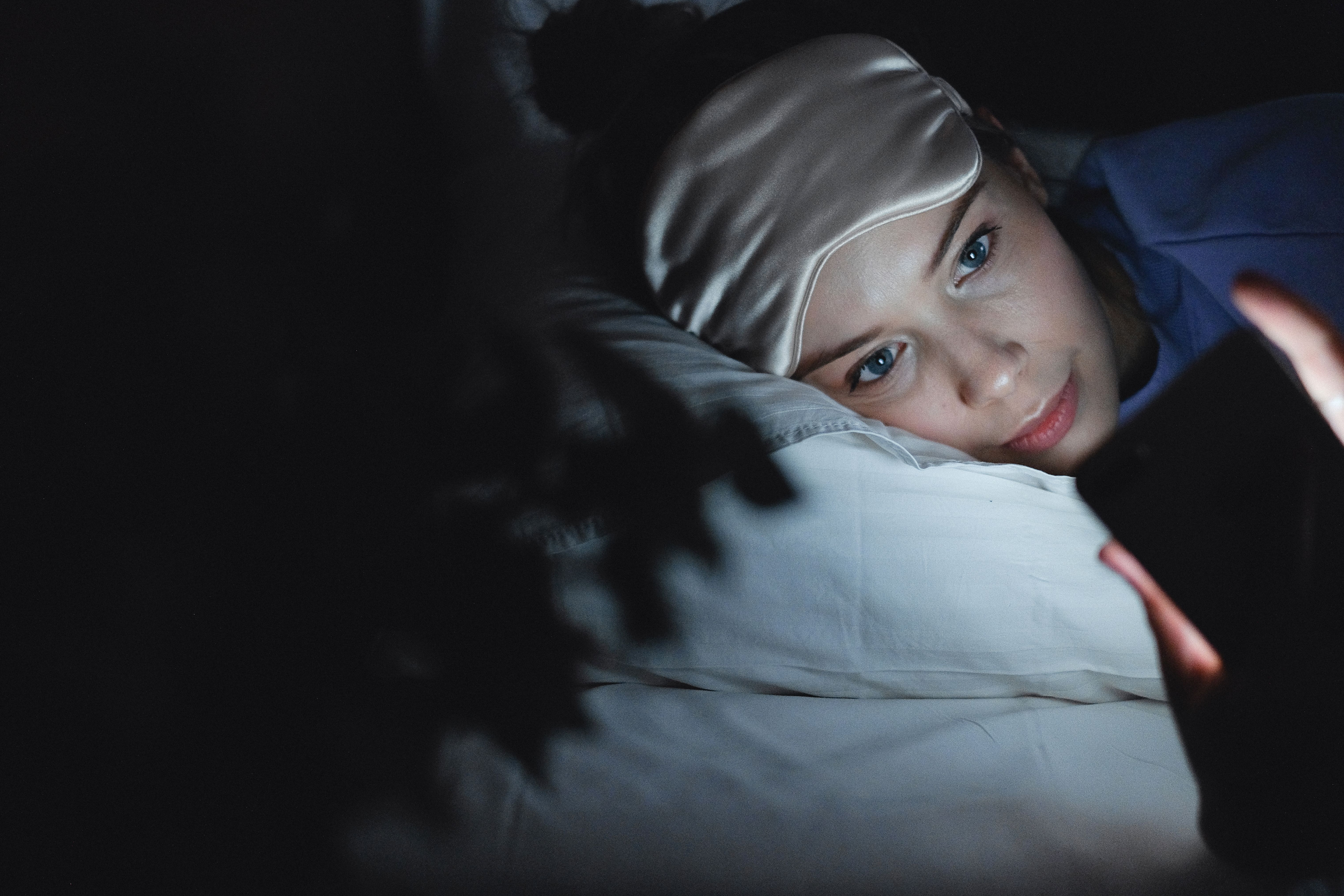 A woman in bed in a dark room, looking at her phone with a sleep mask on her forehead | Source: Pexels