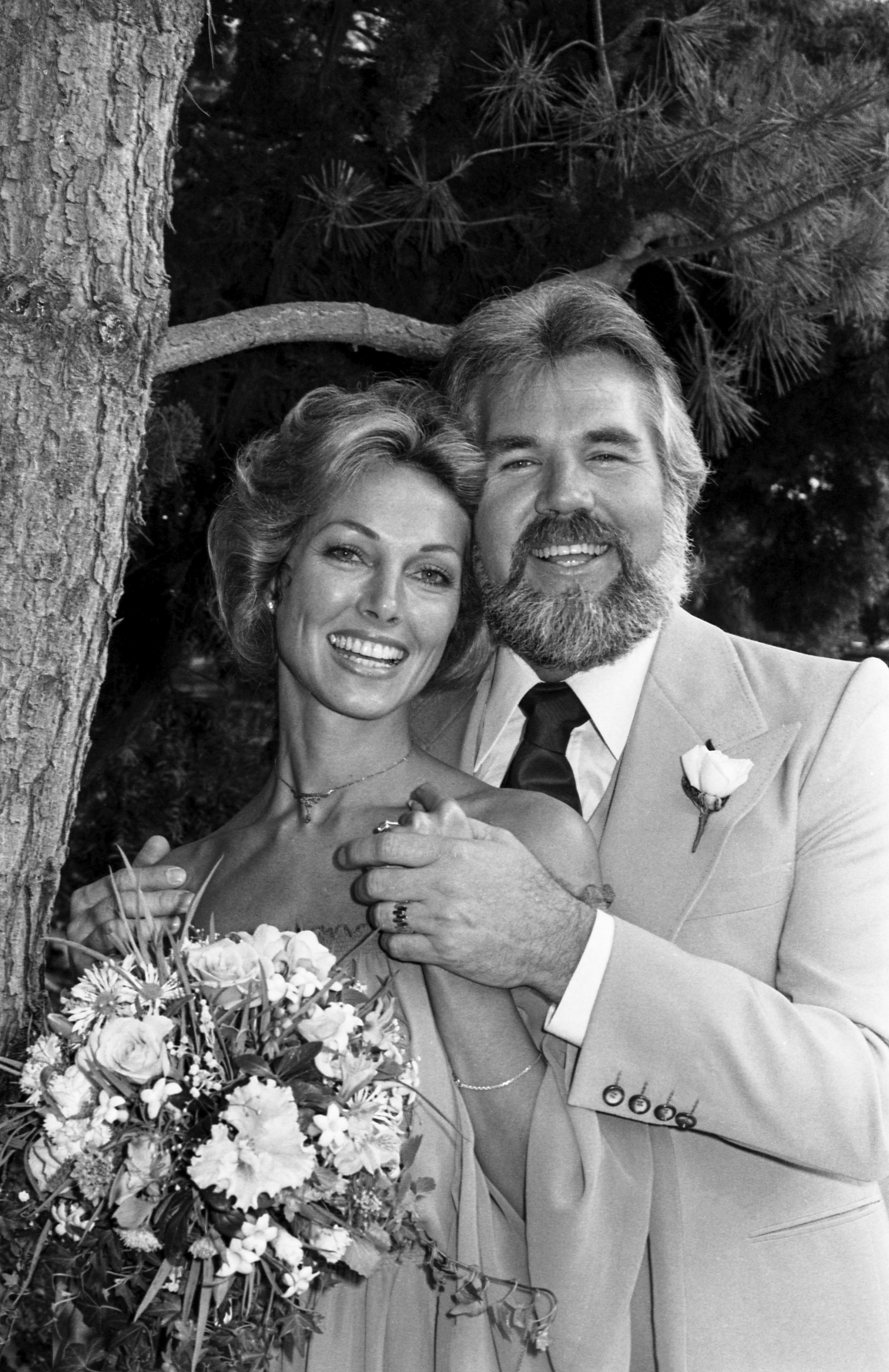 Kenny Rogers and actress Marianne Gordon radiate joy following their wedding ceremony at the singer's home in 1977. Standing together outdoors beneath tree branches, Kenny embraces his fourth wife from behind in a tailored suit, necktie, and white boutonnière, while Marianne holds her bridal floral bouquet and clasps his hands, beaming in a strapless gown and delicate pendant necklace. | Source: Getty Images