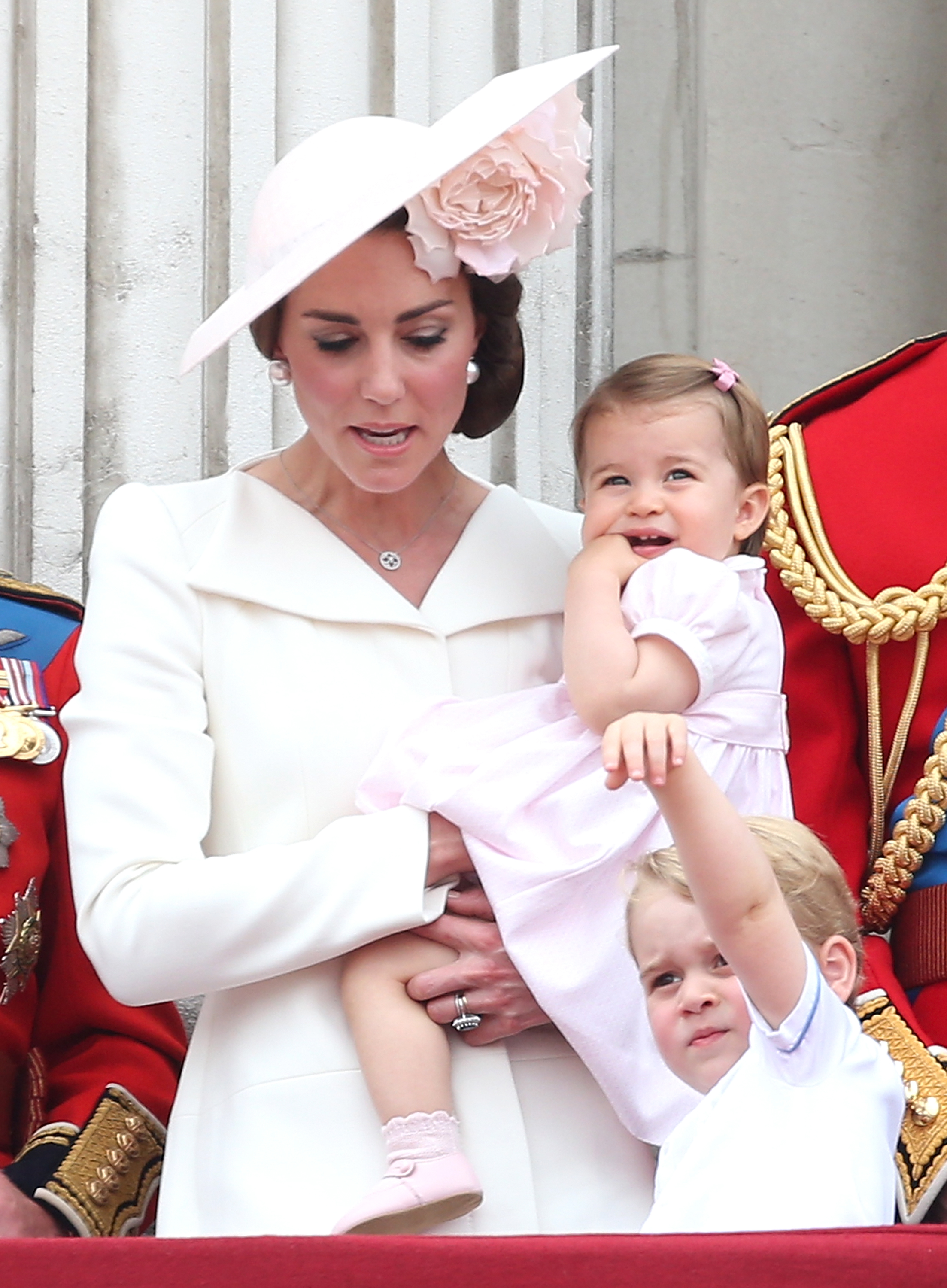 Catherine, Duchess of Cambridge holds a young Princess Charlotte in her arms while standing on the balcony alongside Prince George during the Trooping the Colour parade at The Mall on June 11, 2016, in London, England. Catherine looks radiant in an ivory tailored coat dress and a pale pink Philip Treacy fascinator accented with a large floral blossom, while Charlotte wears a pastel pink puffed-sleeve dress with matching shoes and George reaches upward in a white collared ensemble. | Source: Getty Images