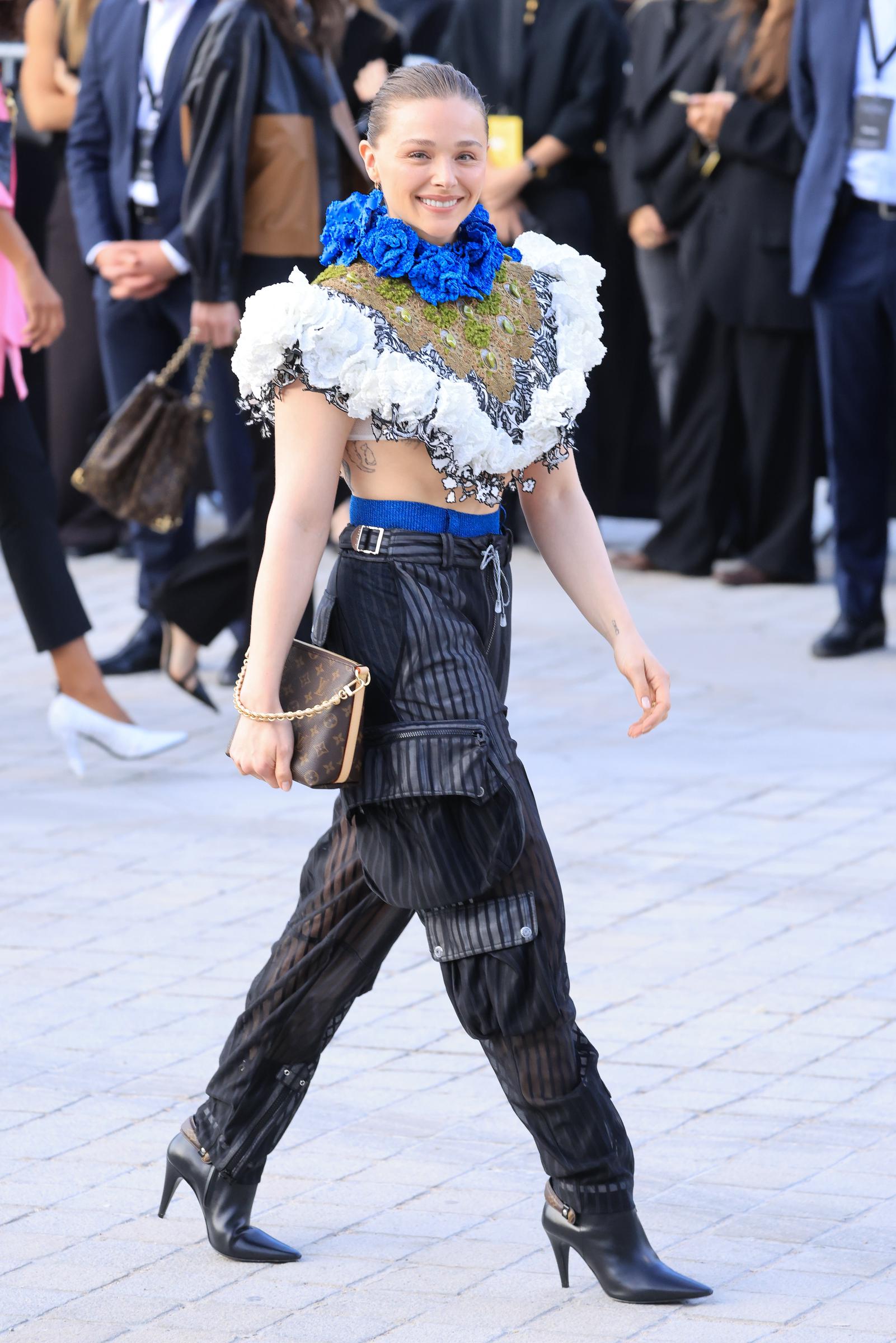 Chloë Grace Moretz attends the Louis Vuitton Womenswear Spring/Summer 2027 show as part of Paris Fashion Week on October 06, 2026, in Paris, France. She wore a dramatic cropped floral top with oversized white appliqués and a vivid blue flower-covered neckline, paired with sheer black cargo trousers and a bright blue belt. Pointed black ankle boots and a classic Louis Vuitton monogram bag completed the bold, texture-heavy look. | Source: Getty Images