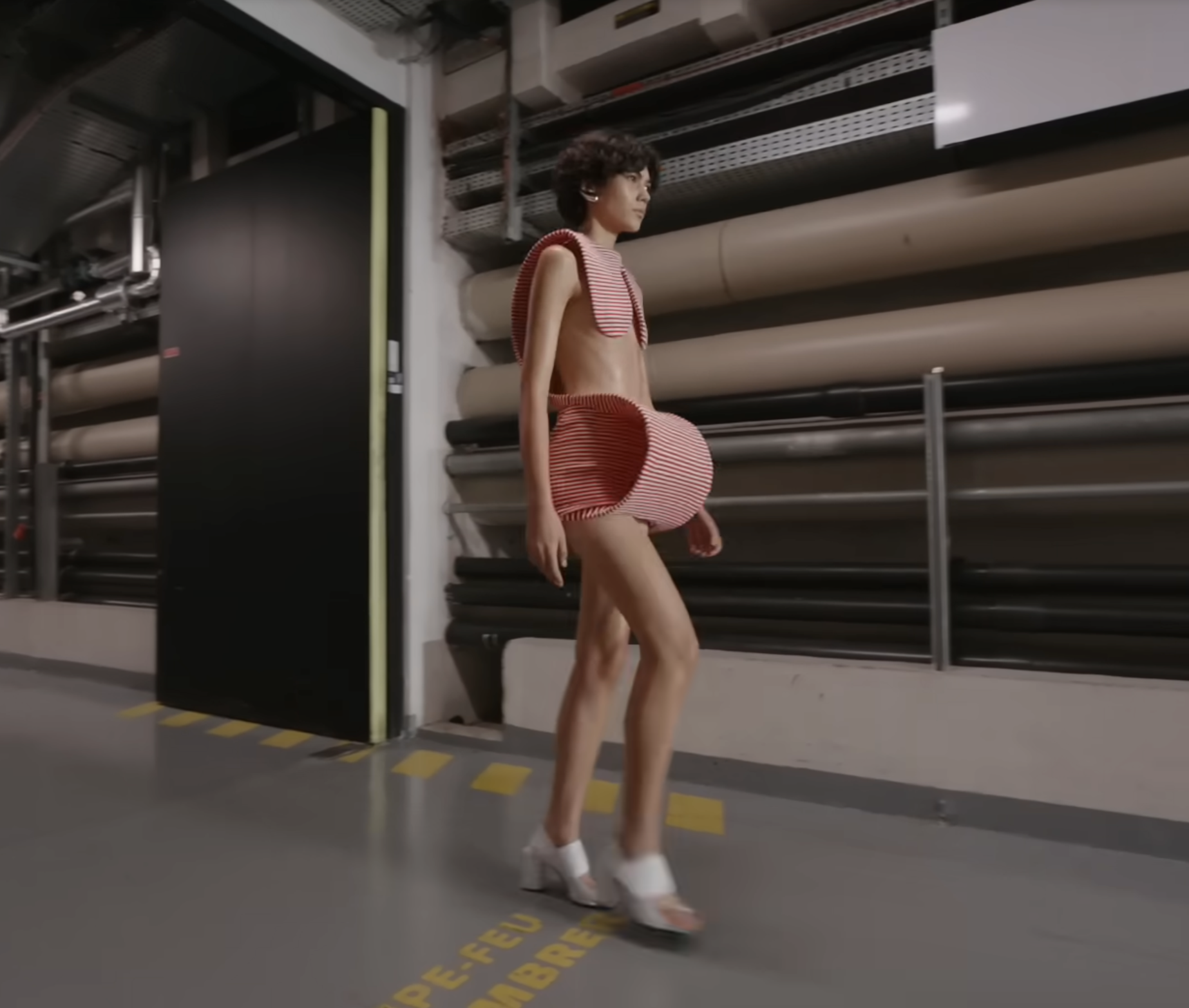 Another standout design is worn by this model, styled with white block heels. The top appears to rest lightly on his shoulders, leaving part of the upper body exposed, while the skirt features a uniquely structured, sculptural build at the front. From a post dated October 5, 2025. Source: Youtube/@jeanpaulgaultier