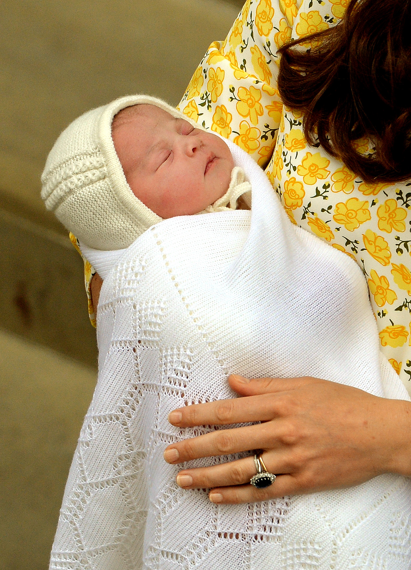 Catherine, Duchess of Cambridge cradles her newborn daughter, Princess Charlotte, outside the Lindo Wing at St Mary's Hospital on May 2, 2015, in London, England. The sleeping infant is swaddled in a soft white knit blanket with a matching bonnet, held securely by Catherine, who wears a yellow floral-print dress and her sapphire engagement ring. | Source: Getty Images