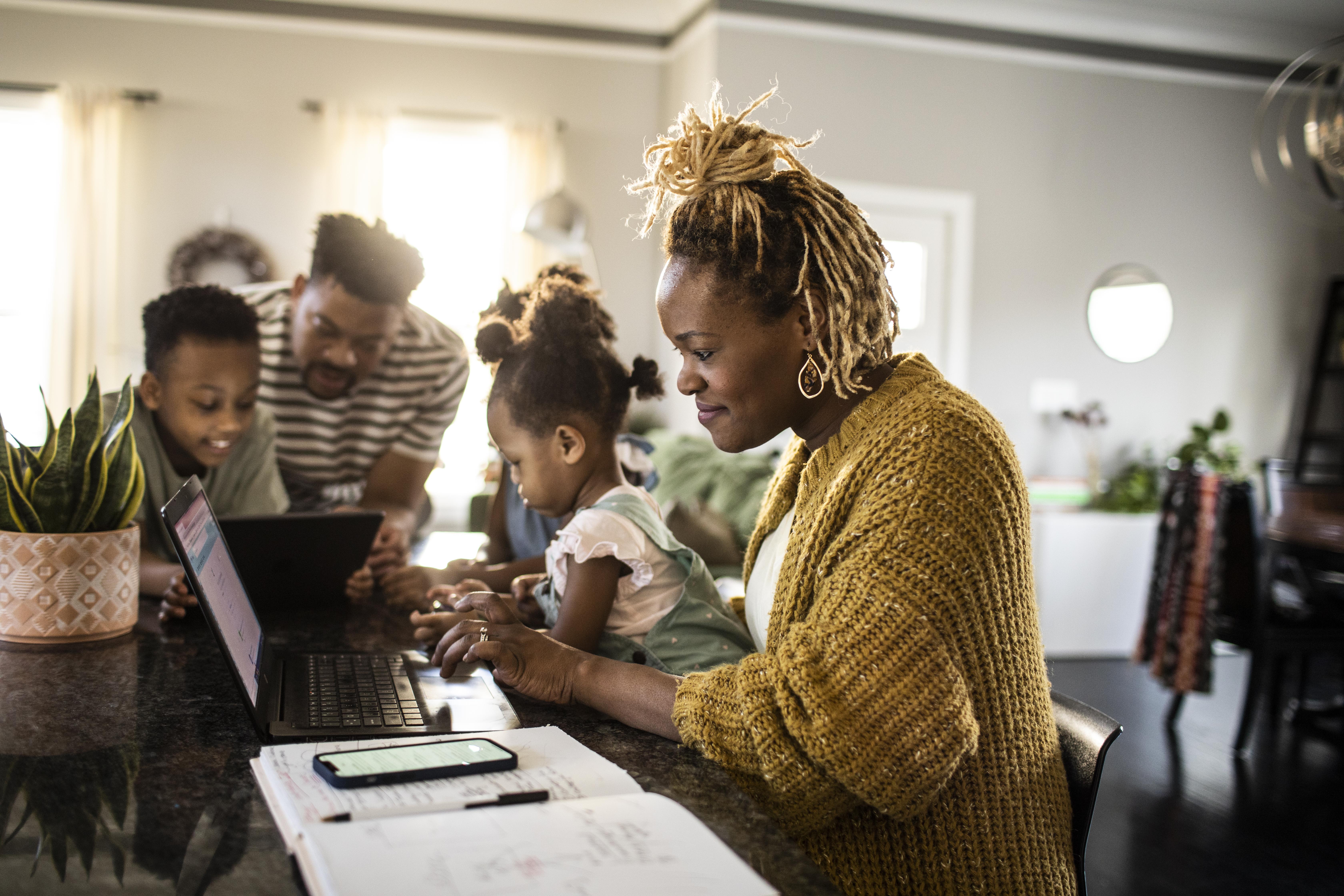 Mother working from home while holding toddler, family in background | Source: Getty Images