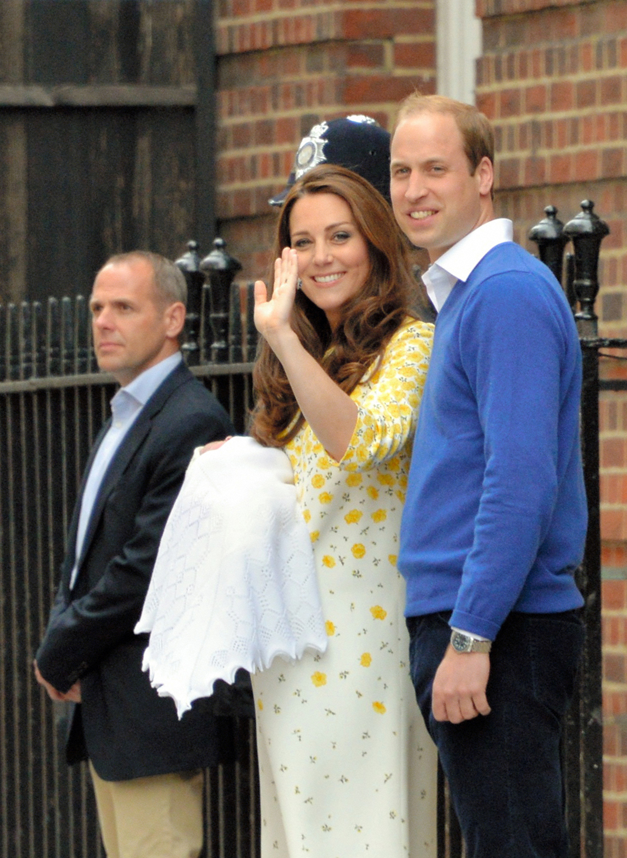 Prince William, Duke of Cambridge smiles proudly alongside Catherine, Duchess of Cambridge as she waves to the cheering crowd gathered outside the steps of the medical facility. William coordinates in a bright royal blue V-neck sweater layered over an open-collar white button-down and dark navy trousers, standing near security as the couple introduces their newly arrived royal family member to the public. | Source: Getty Images