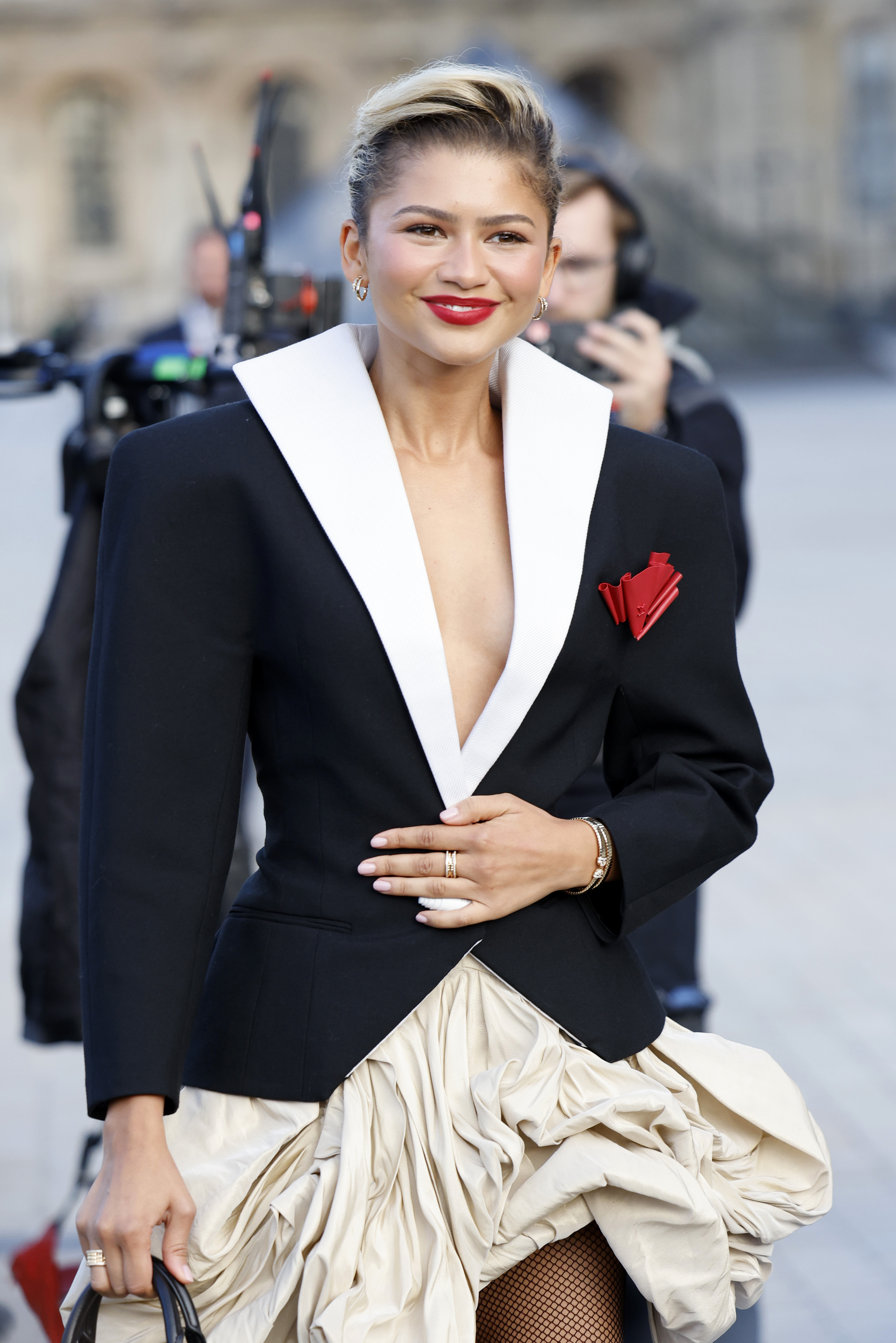 Zendaya attended the Women’s Spring/Summer 2025 show at Paris Fashion Week in France, wearing a sculpted black plunging blazer with exaggerated shoulders and a high white collar. The blazer, fastened with a single white cylindrical button, featured a red rose-like appliqué. She layered it over a beige bubble skirt.