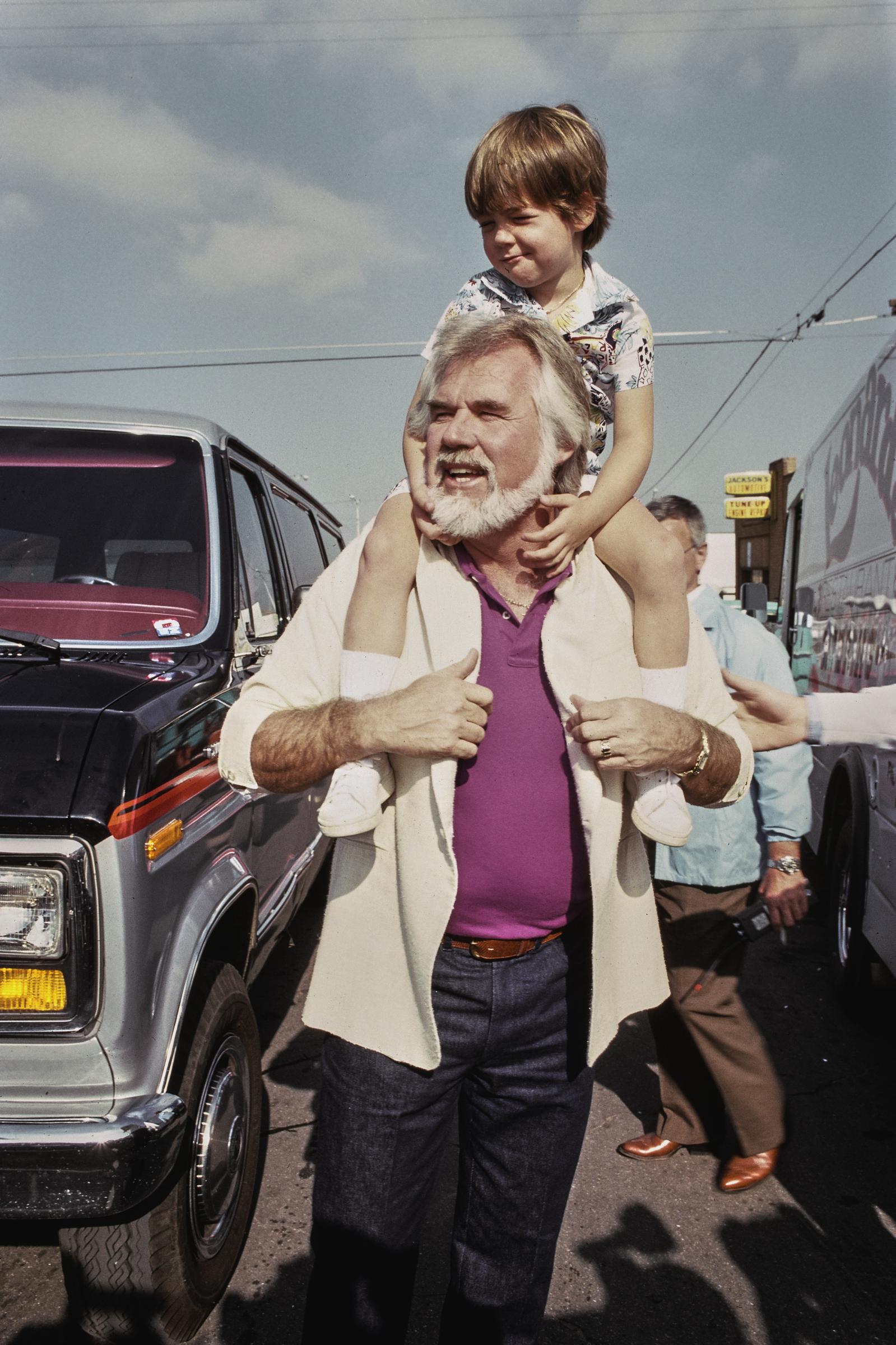 Kenny Rogers carries his young son Christopher Cody Rogers on his shoulders during the commercial and video shoot for the "Hands Across America" campaign on January 18, 1986, in Taft, California. Dressed in an open cream blazer over a purple polo shirt and dark jeans, the musician smiles outdoors as little Christopher rests comfortably from above in a patterned shirt, white socks, and sneakers.