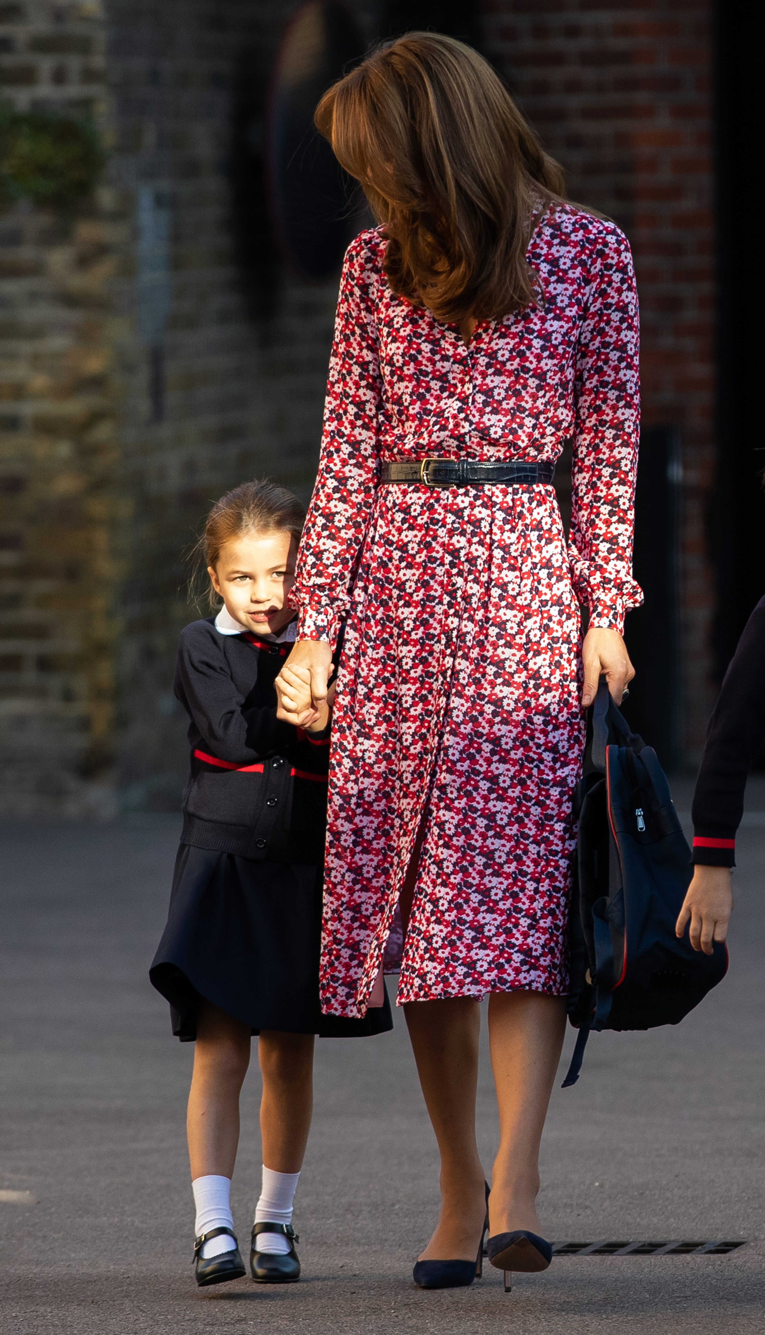 Princess Charlotte holds her mother Catherine's hand as she arrives for her very first day of school at Thomas's Battersea on September 5, 2019, in London, England. The young royal peers shyly toward onlookers in her navy-and-red school uniform skirt, cardigan, white ankle socks, and black Mary Jane shoes, accompanied by the Duchess in a belted pink-and-navy floral midi dress and pointed navy pumps. | Source: Getty Images