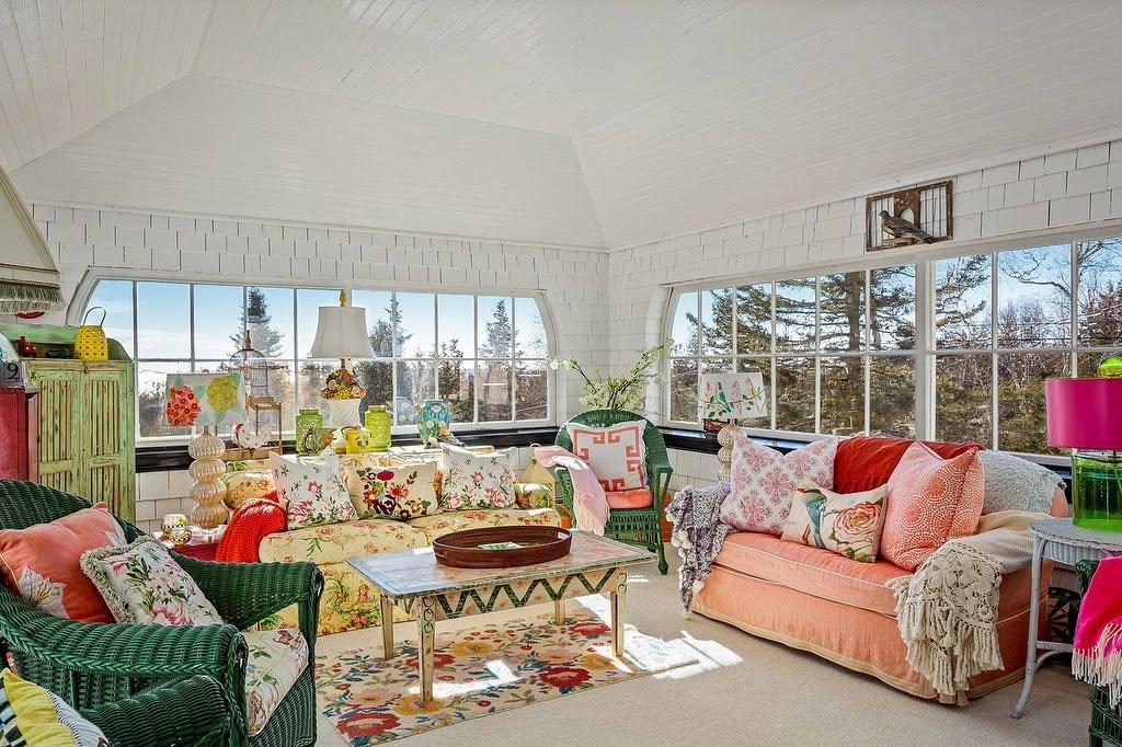 Large windows fill this cheerful sitting room with natural light, while floral patterns, wicker furniture, and colorful textiles enliven the space. | Source: Facebook/LegacySIR