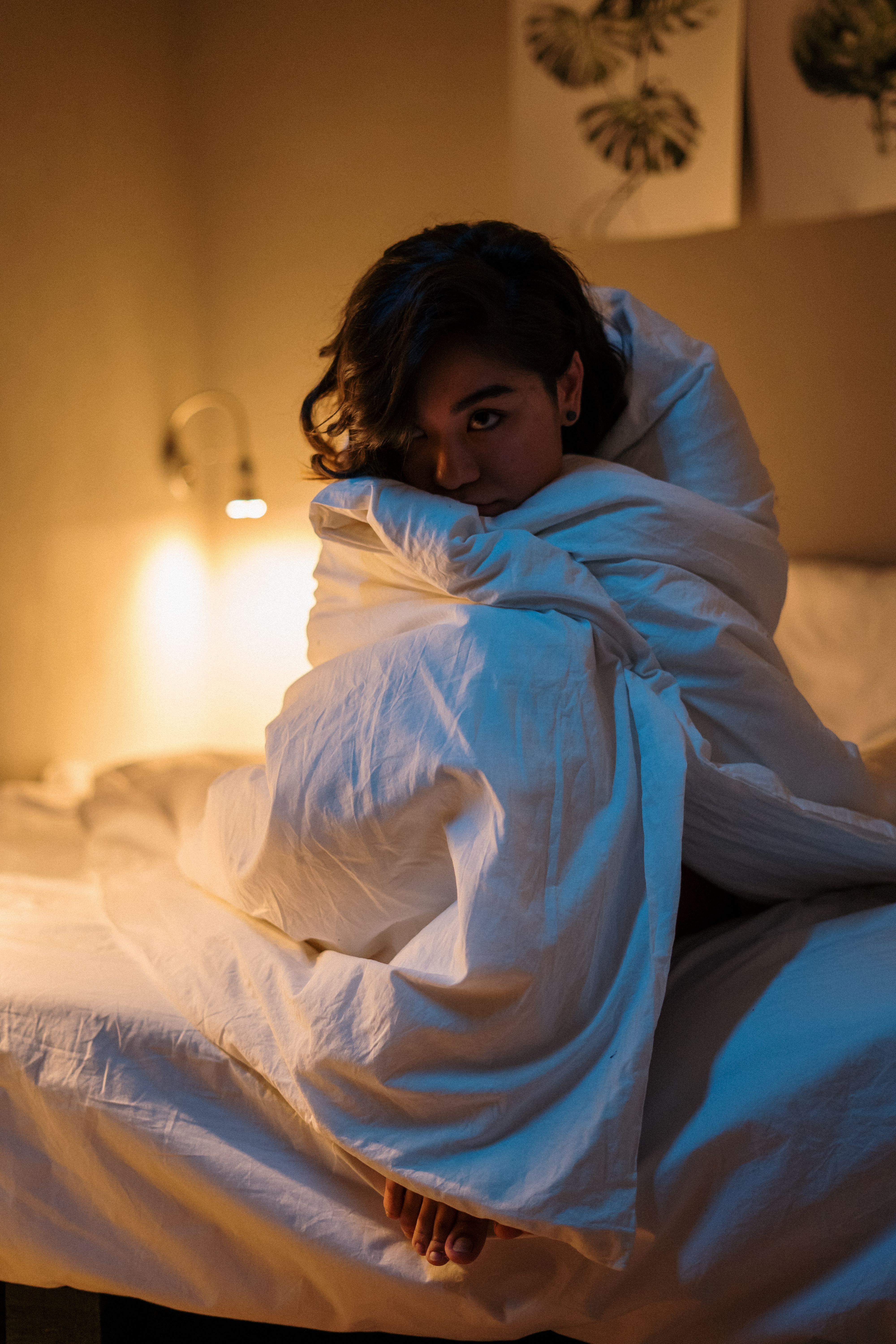 A woman wrapped in a blanket while sitting up in bed at night with a lamp on | Source: Pexels