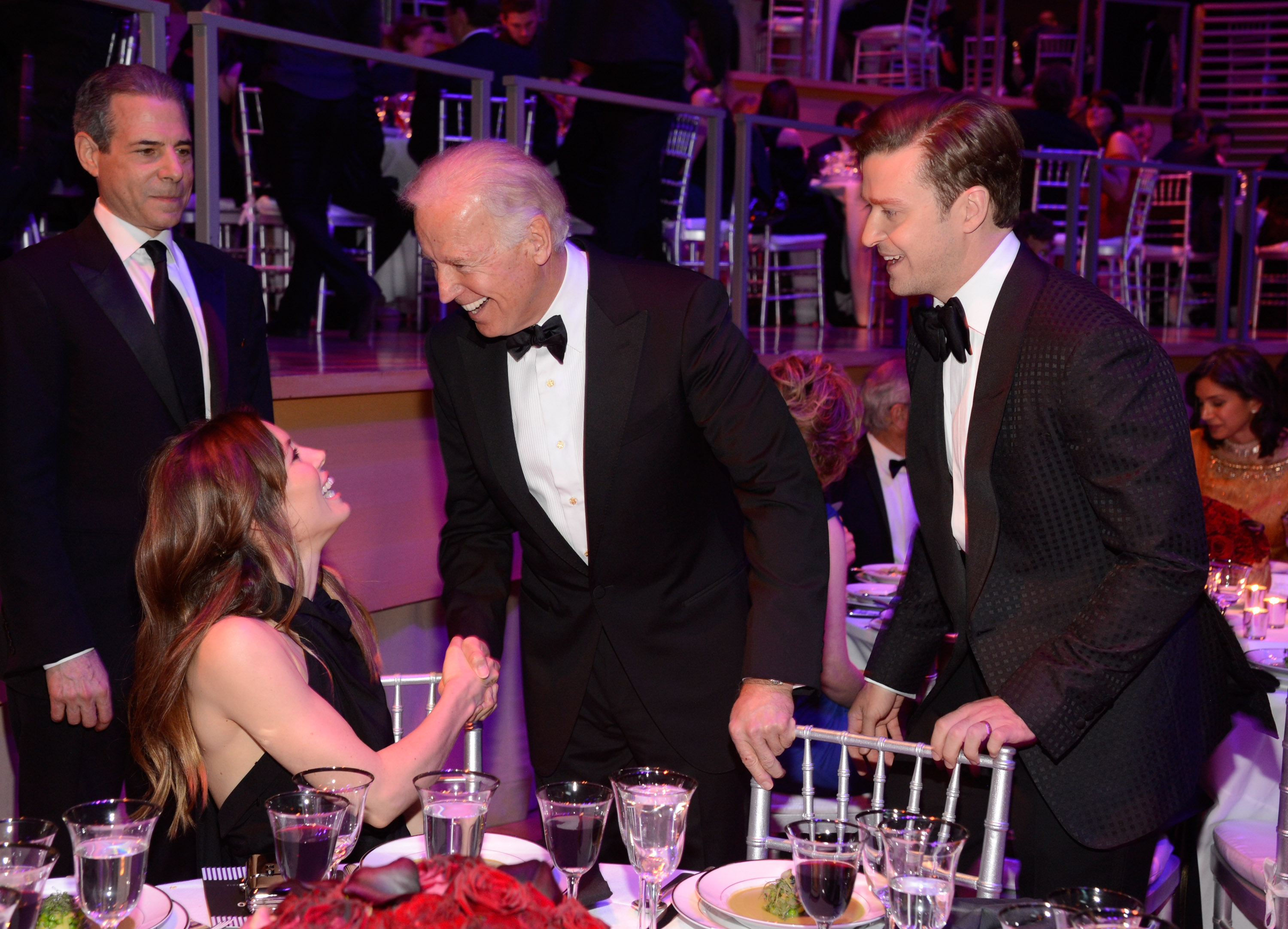 Jessica Biel laughs while greeting former U.S. President Joe Biden at a formal dinner, as Justin Timberlake stands beside them smiling. Timberlake wears a black patterned tuxedo, while Biel is dressed in a sleeveless black outfit.
