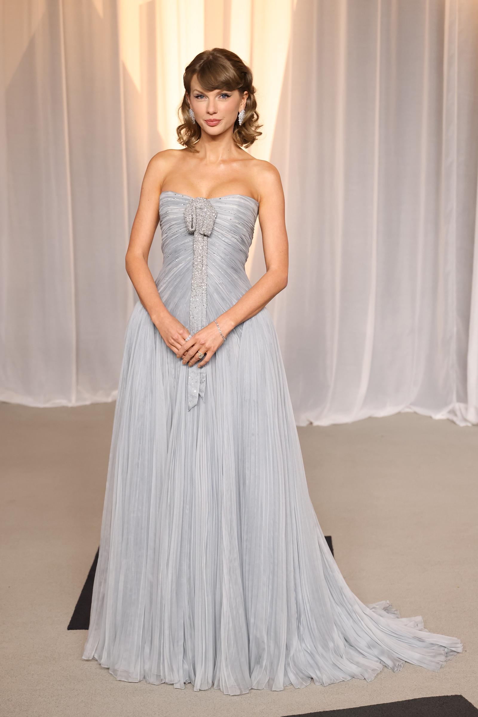 A full-length view highlights the gown's fitted, ruched bodice, voluminous pleated skirt, and long jeweled detail extending from the neckline.