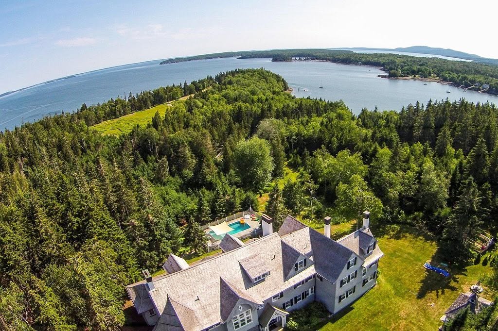 An aerial view shows the expansive home surrounded by dense woodland, with its oceanfront setting and nearby waters stretching into the distance. | Source: Facebook/LegacySIR