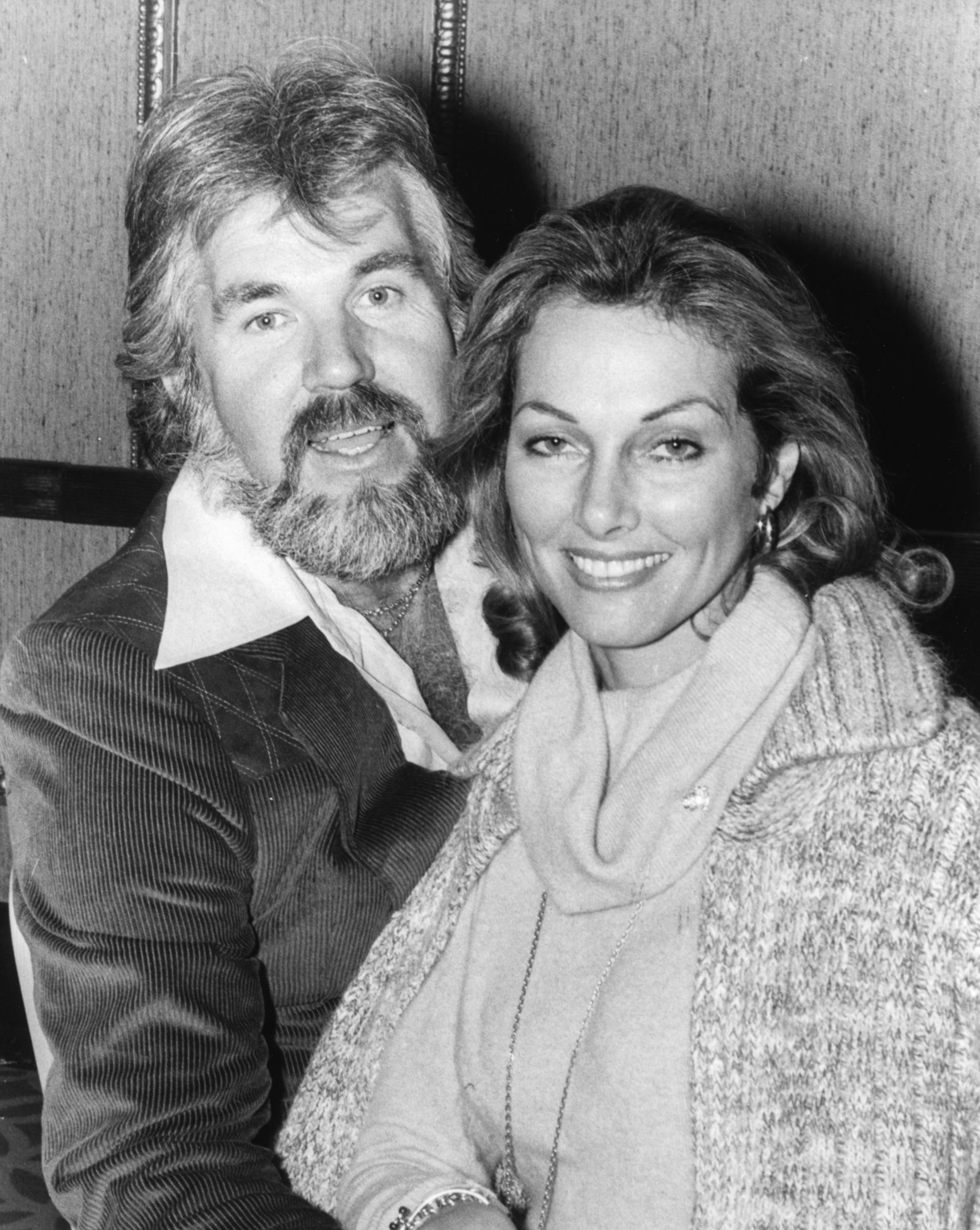 Kenny Rogers and his fourth wife, actress Marianne Gordon, pose for a portrait following their arrival in Britain ahead of his concert tour on November 7, 1977. Married earlier that same year, the couple smile warmly together—Kenny in a corduroy jacket and wide-collared shirt, and Marianne in a soft cowl-neck sweater paired with a cozy knit cardigan and delicate jewelry.