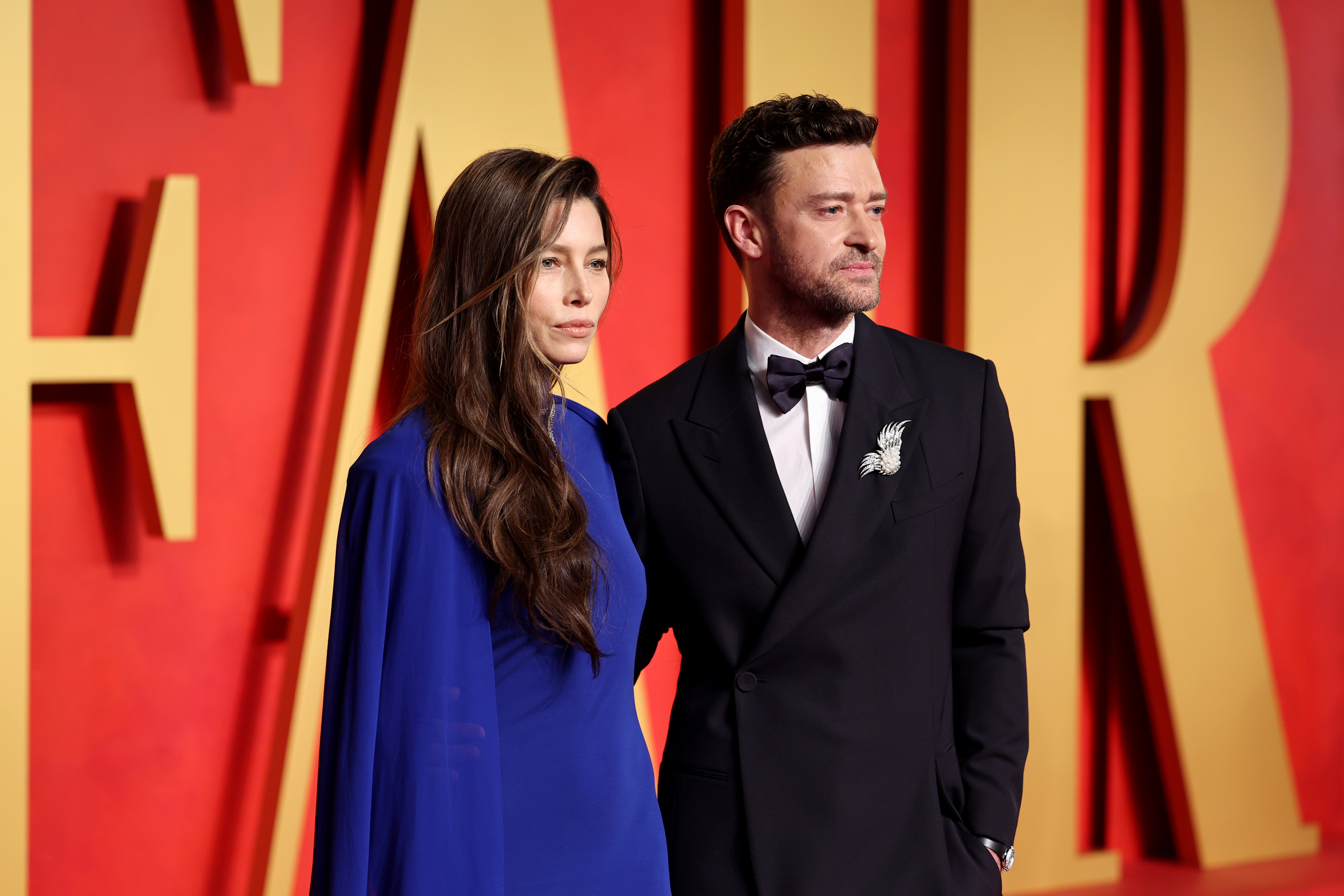 Jessica Biel and Justin Timberlake pose together on a red carpet. Biel wears a striking royal-blue gown with a flowing silhouette, while Timberlake looks sharp in a black tuxedo with a bow tie and decorative silver brooch.