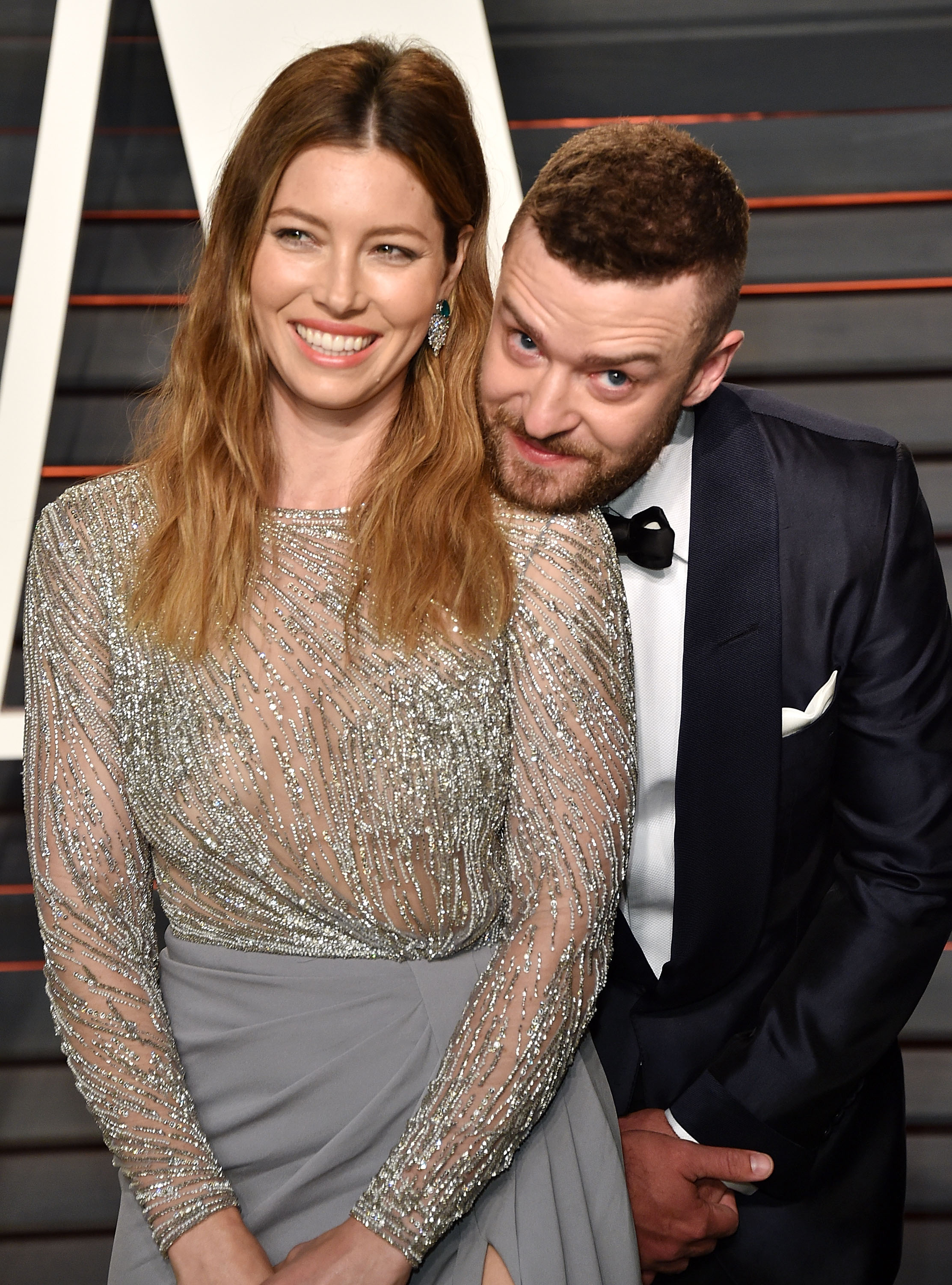 Justin Timberlake leans playfully toward Jessica Biel as they pose together at a glamorous event. Biel smiles in a sheer, silver-beaded top paired with a gray skirt, while Timberlake looks sharp in a dark tuxedo with a bow tie.