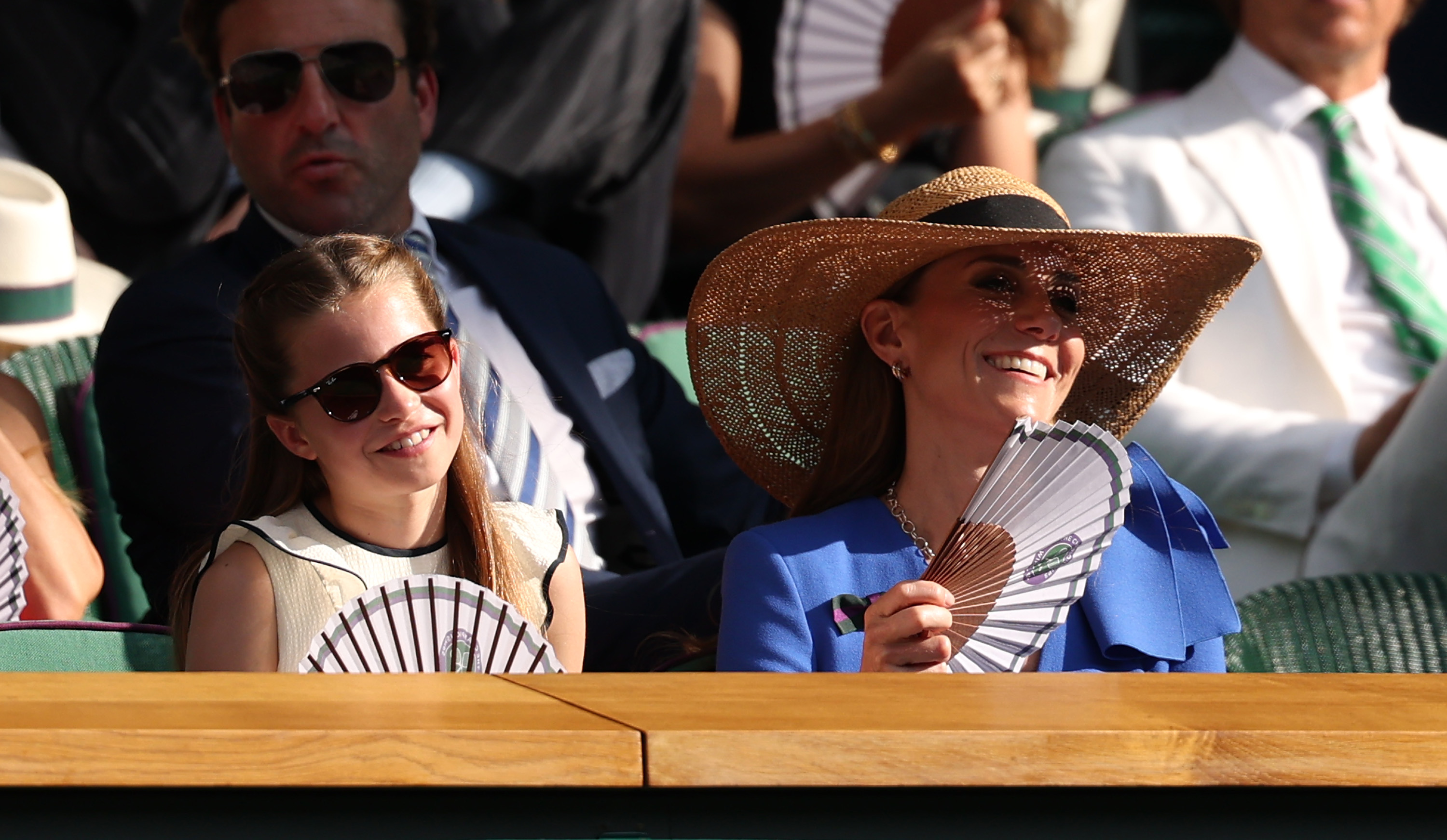 Princess Charlotte of Wales and Catherine, Princess of Wales smile while keeping cool with paper hand fans in the Royal Box on day fourteen of The Championships, Wimbledon, at the All England Lawn Tennis and Croquet Club on July 13, 2025, in London, England. Charlotte watches the Gentlemen's Singles Final in dark tortoiseshell sunglasses and a ruffled cream sleeveless top, sitting beside Catherine, who looks summery in a bright blue dress, statement sunglasses, and a wide-brimmed straw hat. | Source: Getty Images
