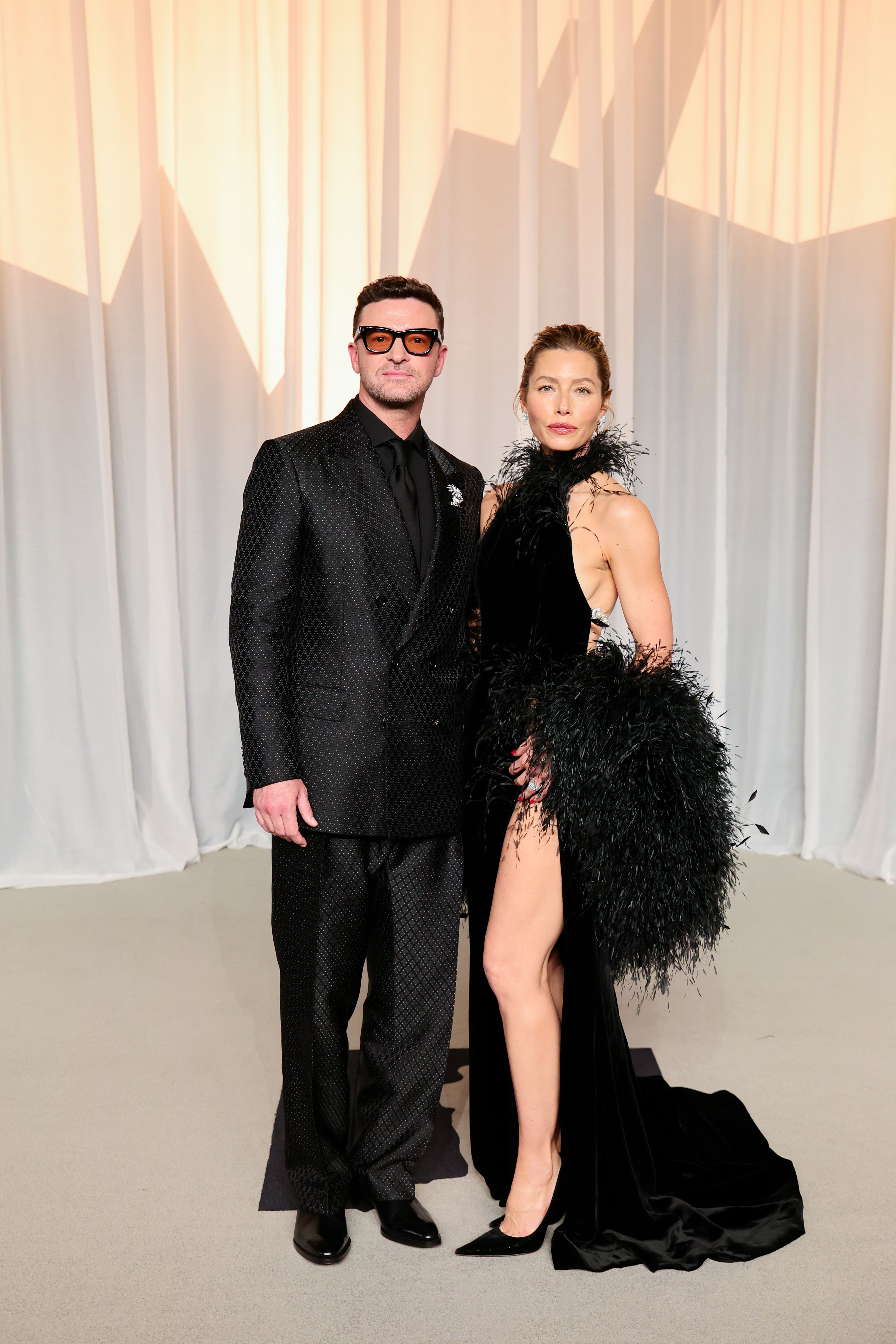 Jessica Biel and Justin Timberlake pose together at a glamorous event. Biel wears a dramatic black gown featuring a feathered neckline and voluminous feathered detailing around the skirt, while Timberlake sports a textured black suit, black shirt, and amber-tinted glasses.