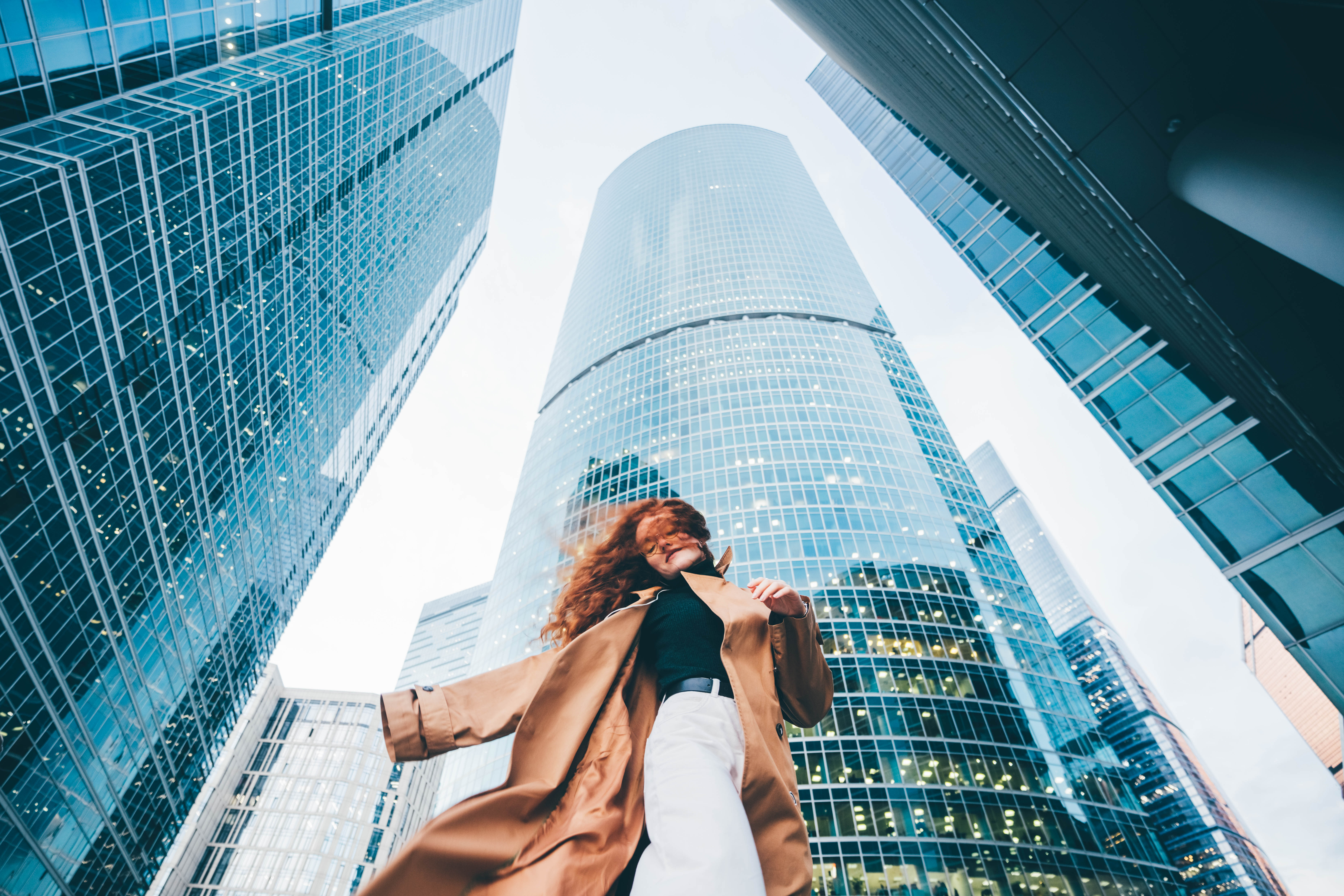 A woman spinning in a brown trench coat amidst towering glass skyscrapers | Source: Getty Images