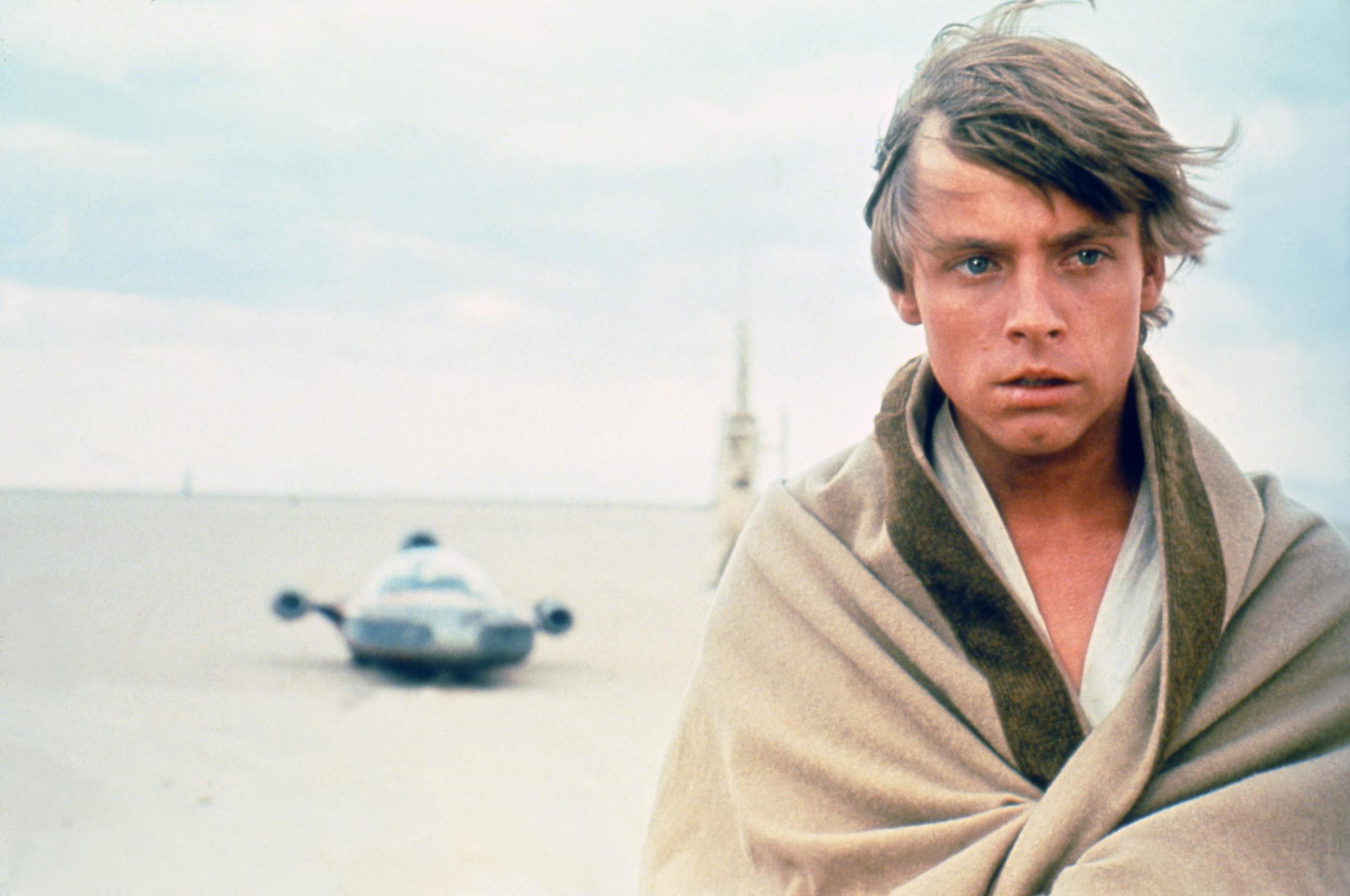 The actor stood against the stark desert landscape during production of "Star Wars: A New Hope," with the character's landspeeder visible in the distance. Wrapped in light-colored clothing beneath the open sky, he embodied the young hero before audiences knew his name. The production still provided another clear view of his appearance before the January 1977 accident that resulted in reconstructive treatment for his facial injuries. | Source: Getty Images