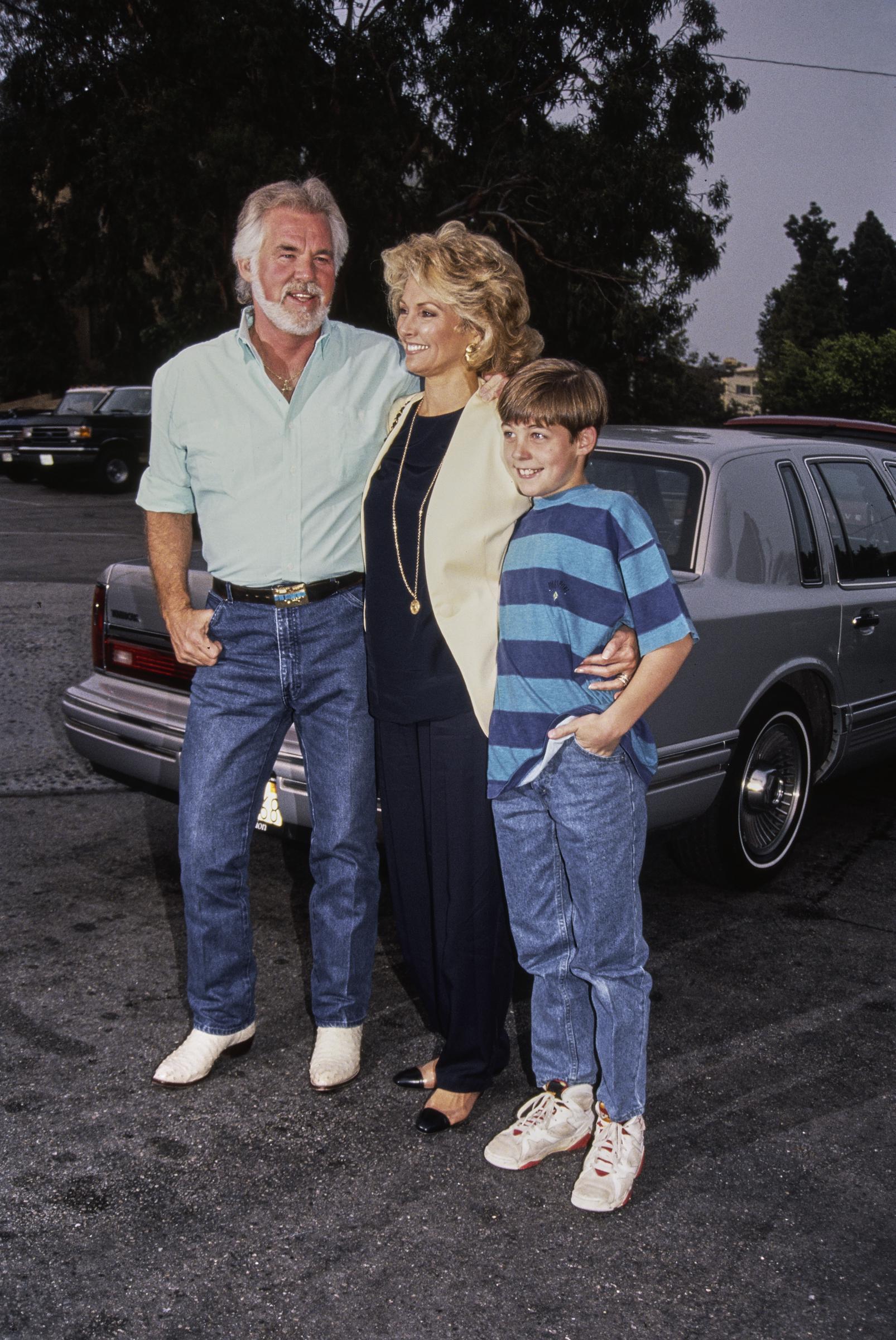 Kenny Rogers poses by a car in Los Angeles alongside his fourth wife, actress Marianne Gordon, and their young son Christopher Cody Rogers circa 1990. The country star stands casually in a mint short-sleeve button-down shirt, blue jeans, and white boots, while Marianne smiles in a black outfit paired with a cream cardigan and gold necklace, resting her hand on Christopher, who smiles in a blue striped T-shirt, jeans, and sneakers.