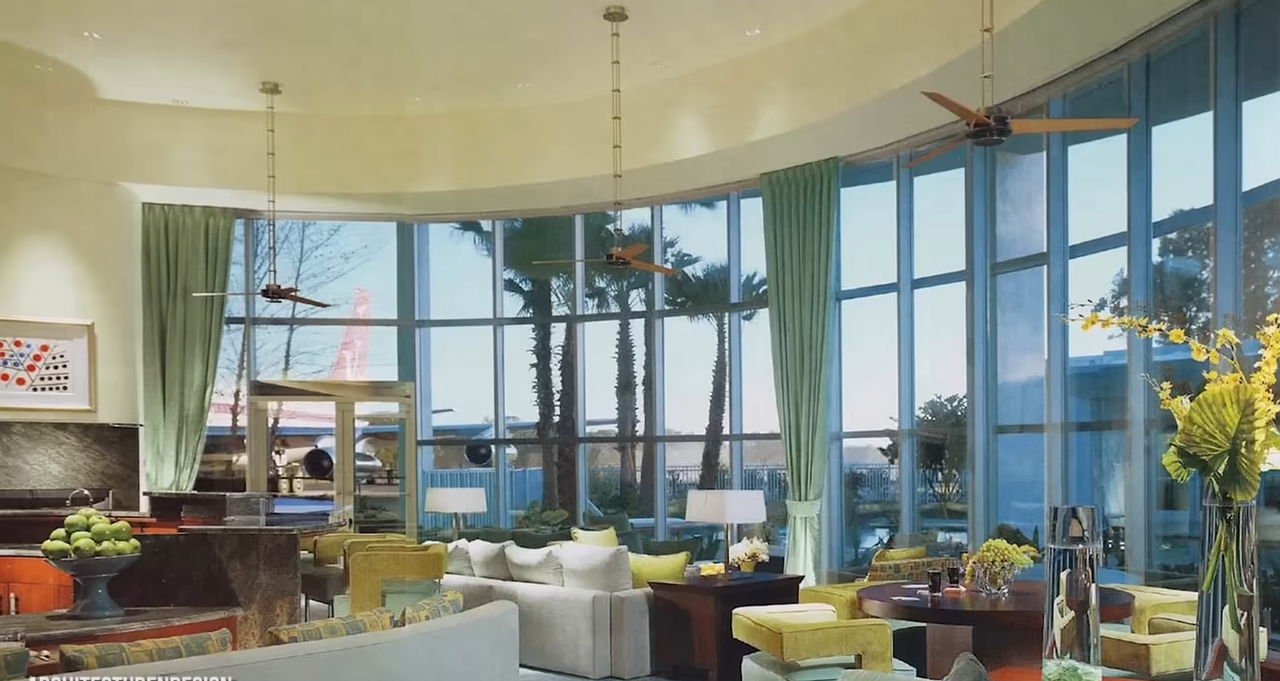 The home's curved living area features soaring windows, pale green curtains, and an unmistakable view of an aircraft outside. | Source: YouTube/@Therichest