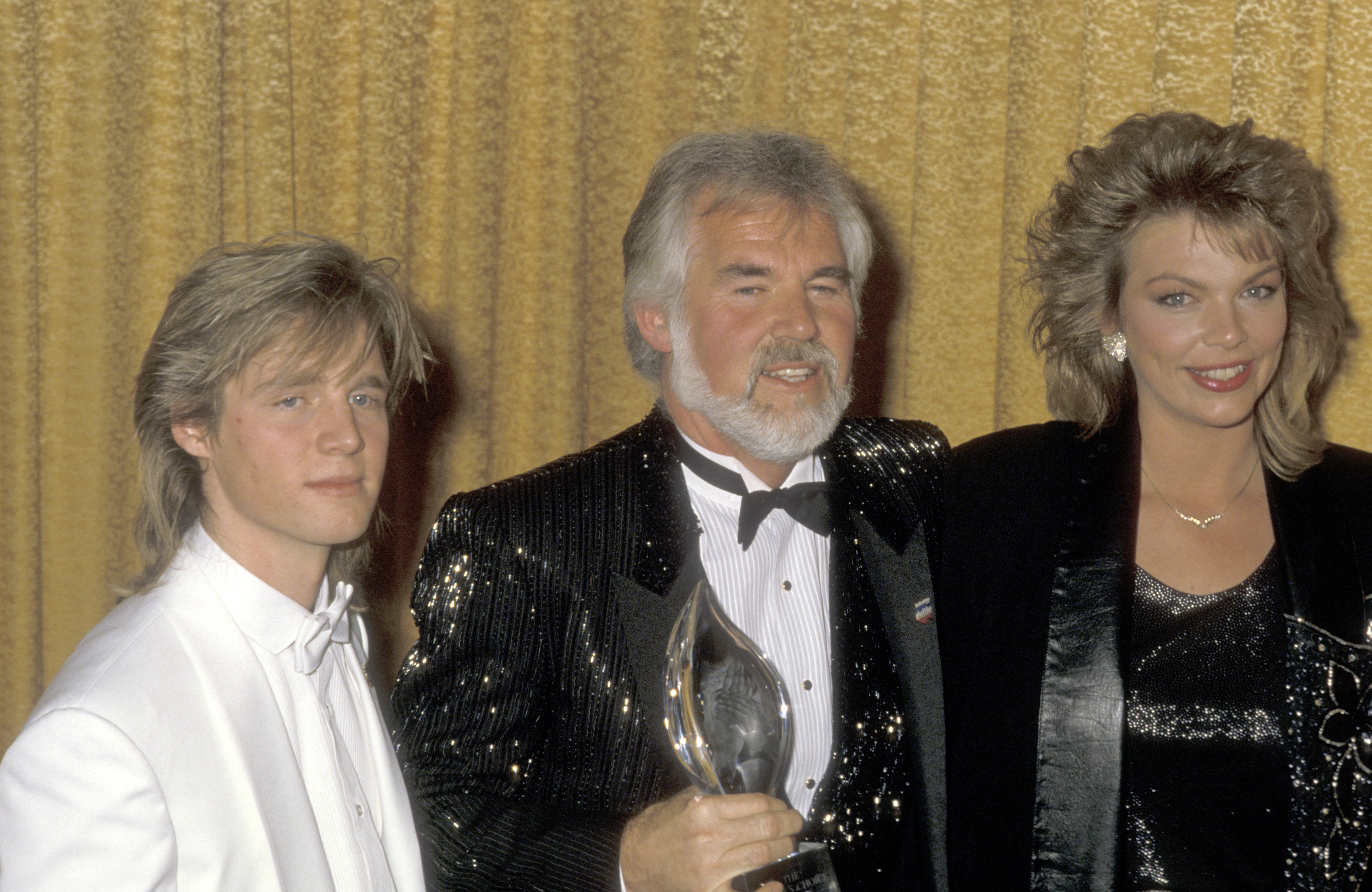 Kenny Rogers celebrates his win at the 12th Annual People's Choice Awards alongside his son Kenny Rogers Jr. and daughter Carole Rogers on March 11, 1986, in Santa Monica, California. Kenny proudly holds his flame-shaped trophy wearing a shimmering sequined tuxedo and bow tie, joined by Kenny Jr. in a crisp white tuxedo jacket and Carole smiling beside them in an embellished black evening jacket. | Source: Getty Images