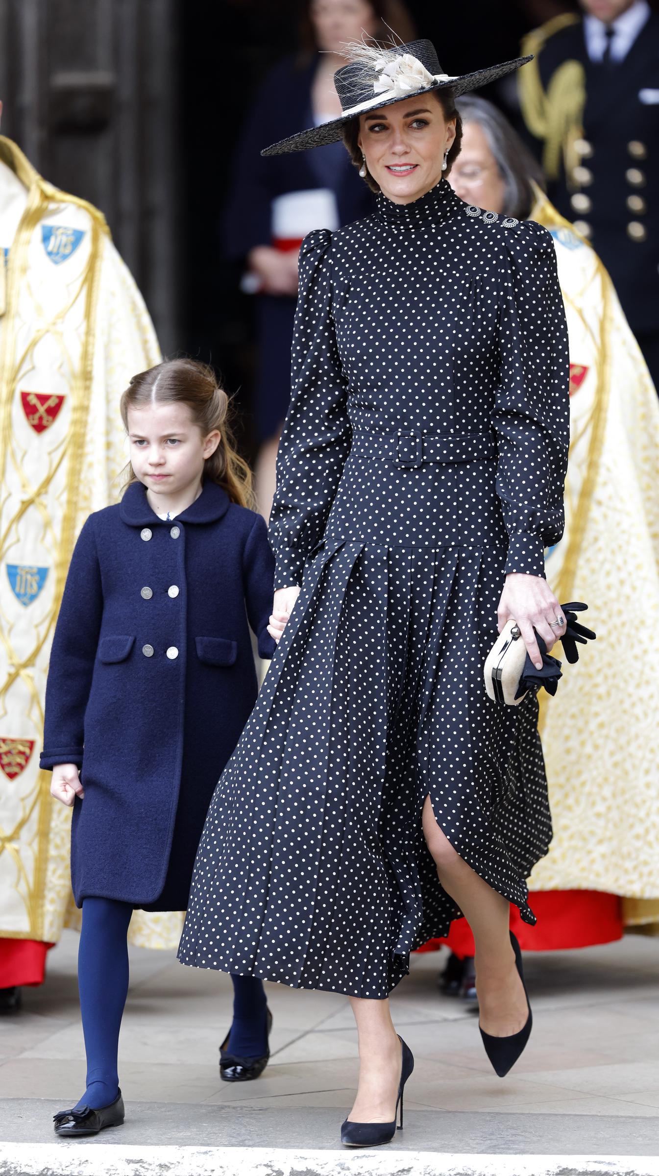 Princess Charlotte walks hand in hand with Catherine, Duchess of Cambridge as they depart Westminster Abbey following the Service of Thanksgiving for the life of Prince Philip, Duke of Edinburgh, on March 29, 2022, in London, England. Charlotte wears a buttoned navy coat over matching tights and black patent shoes, holding hands with Catherine, who is dressed in an elegant black-and-white polka-dot pleated midi dress, wide-brimmed hat with floral accents, and pointed black heels. | Source: Getty Images