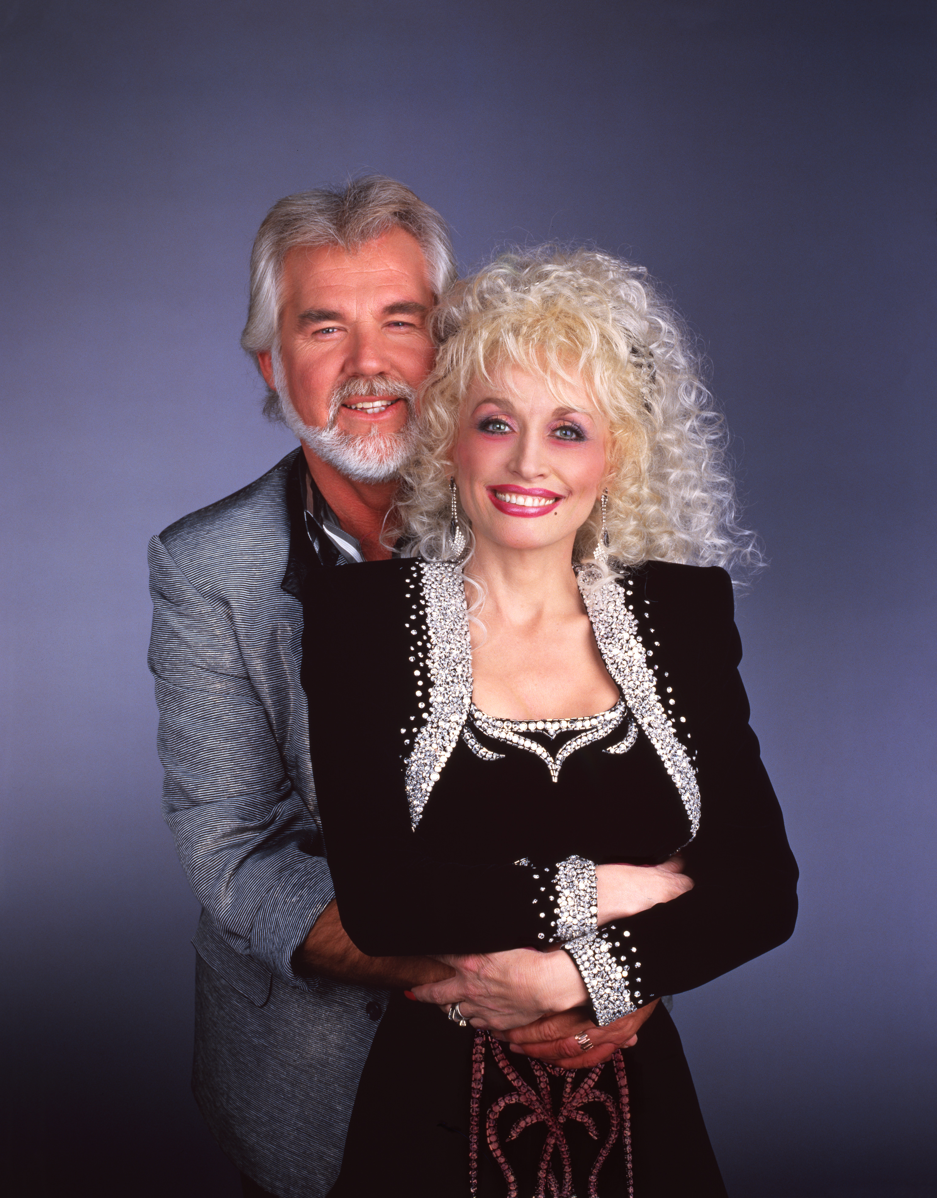 Longtime musical collaborators and close friends Kenny Rogers and Dolly Parton pose for a portrait in 1987. Kenny wraps his arms affectionately around Dolly from behind while wearing a grey textured blazer, as Dolly smiles brightly in an elegant black long-sleeve dress accented with elaborate silver rhinestone embellishments along the neckline, bodice, and cuffs.