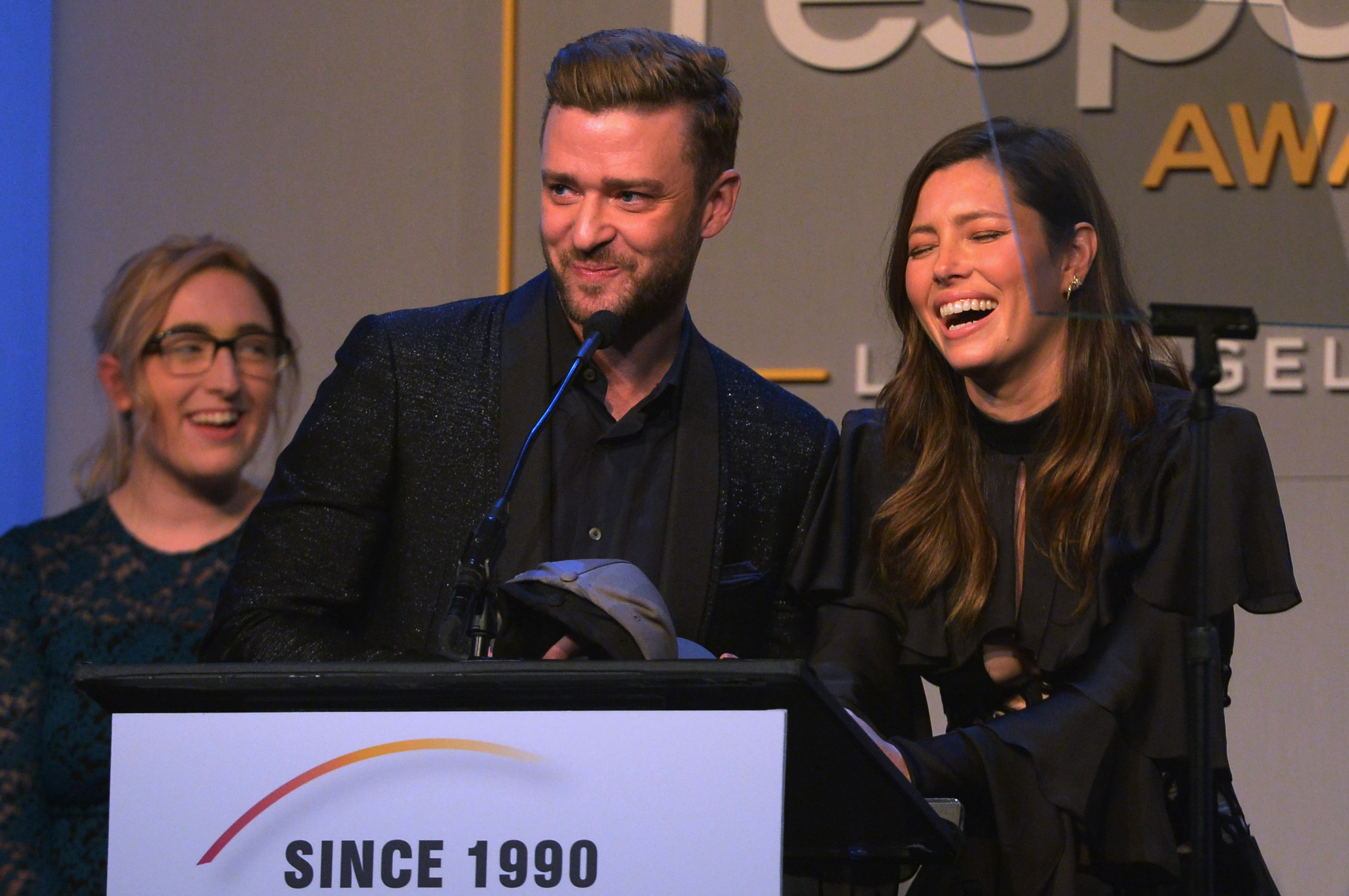 Justin Timberlake and Jessica Biel laugh together at a podium during the GLSEN Respect Awards. Timberlake wears a dark suit, while Biel looks joyful in a black outfit with layered sleeves.